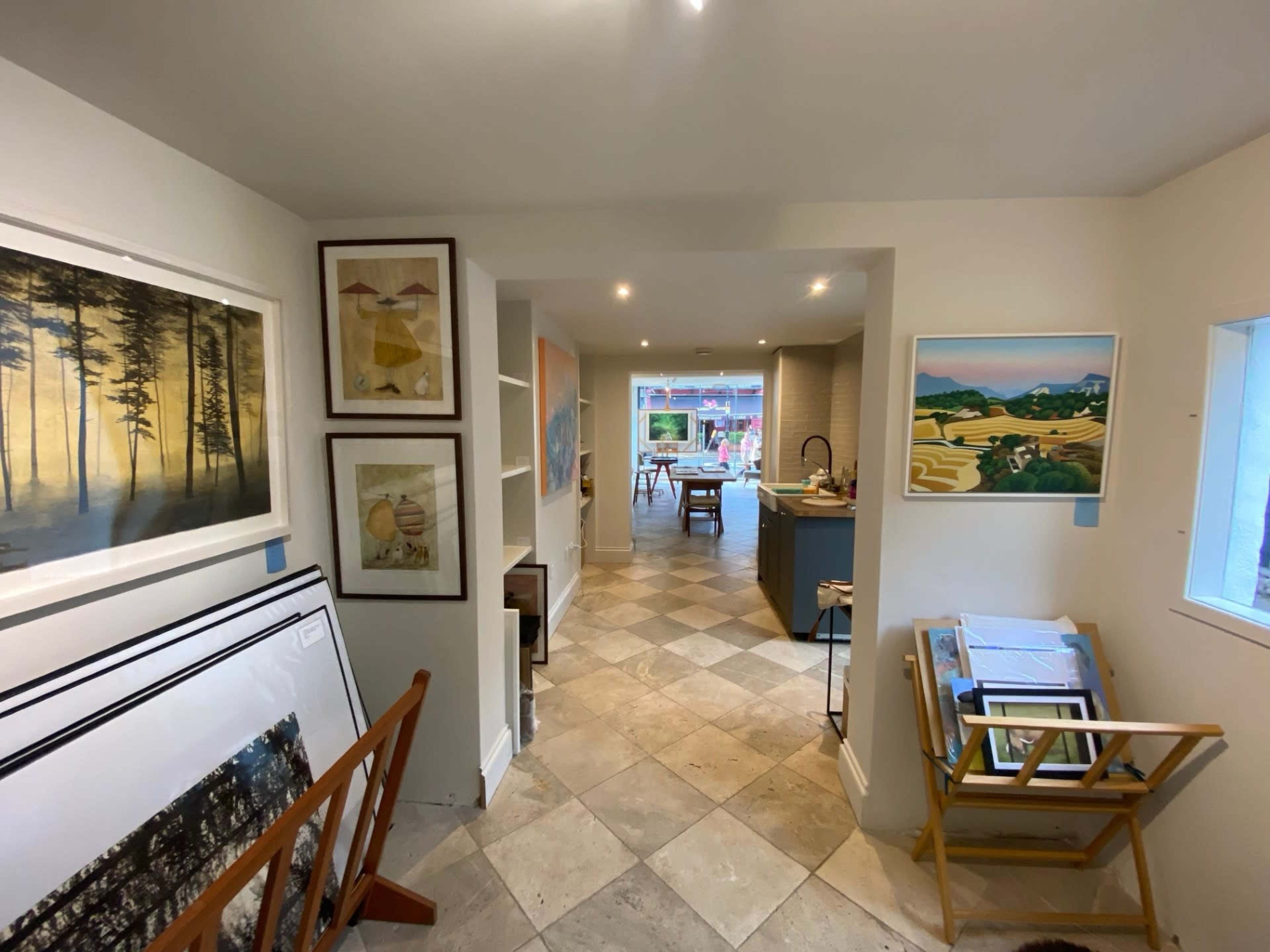 A hallway features framed artwork on the walls, leading to a well-lit open area with a kitchen and additional art displays.