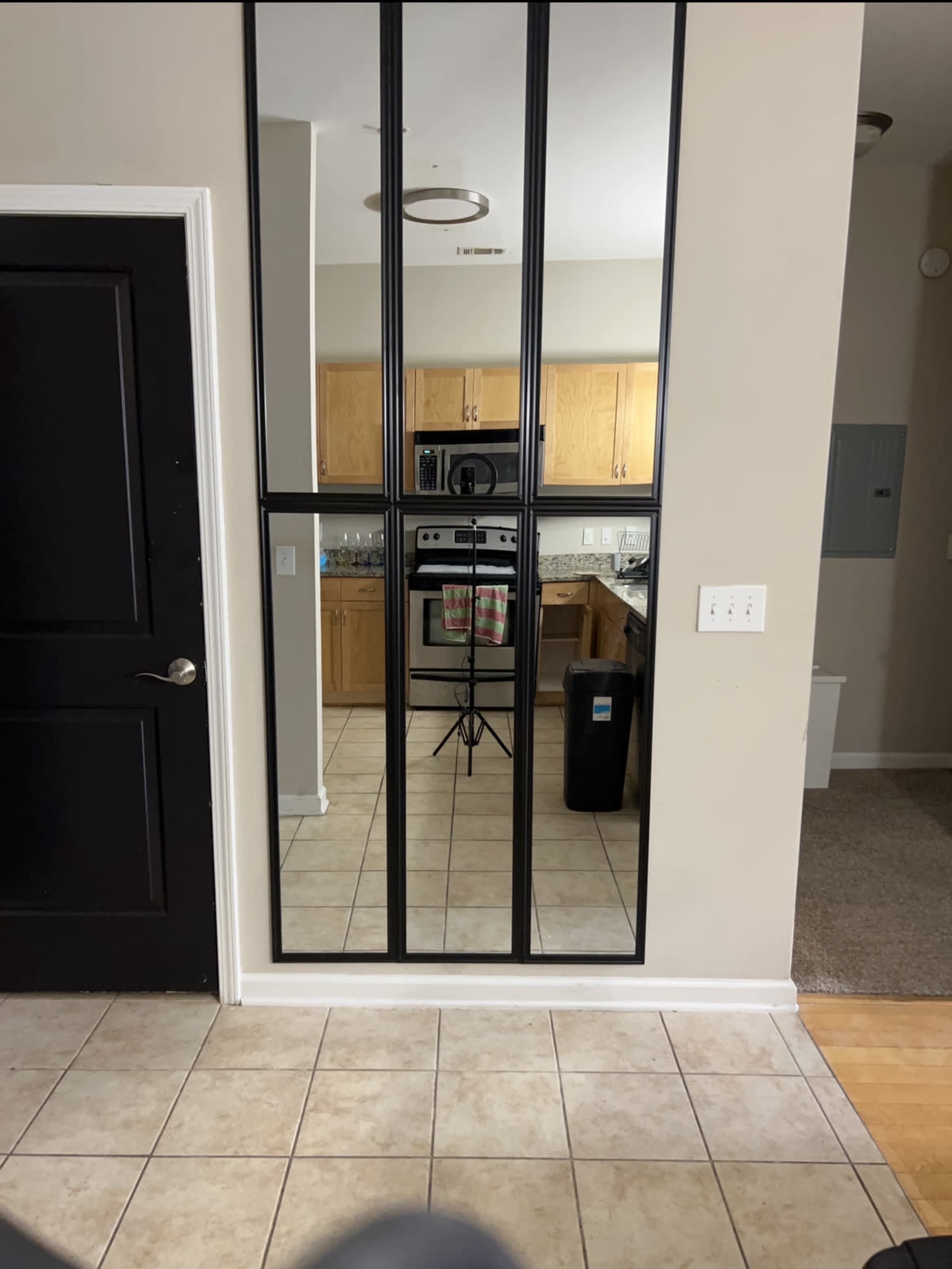A hallway with a large, multi-pane mirror reflects a kitchen that features wooden cabinets and a black oven.