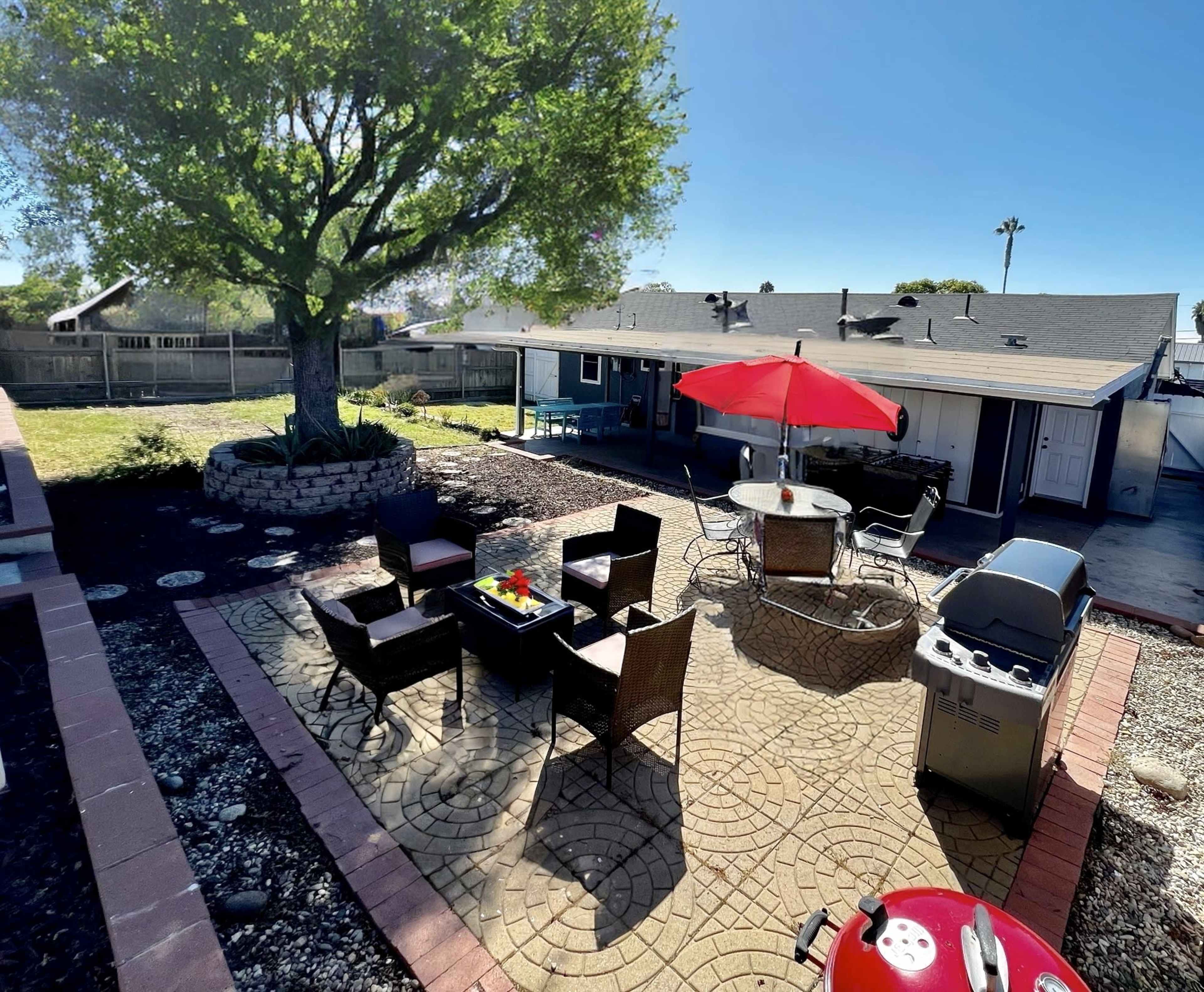 A backyard patio area with a stone pathway, outdoor seating arrangement, a grill, and a large tree providing shade.
