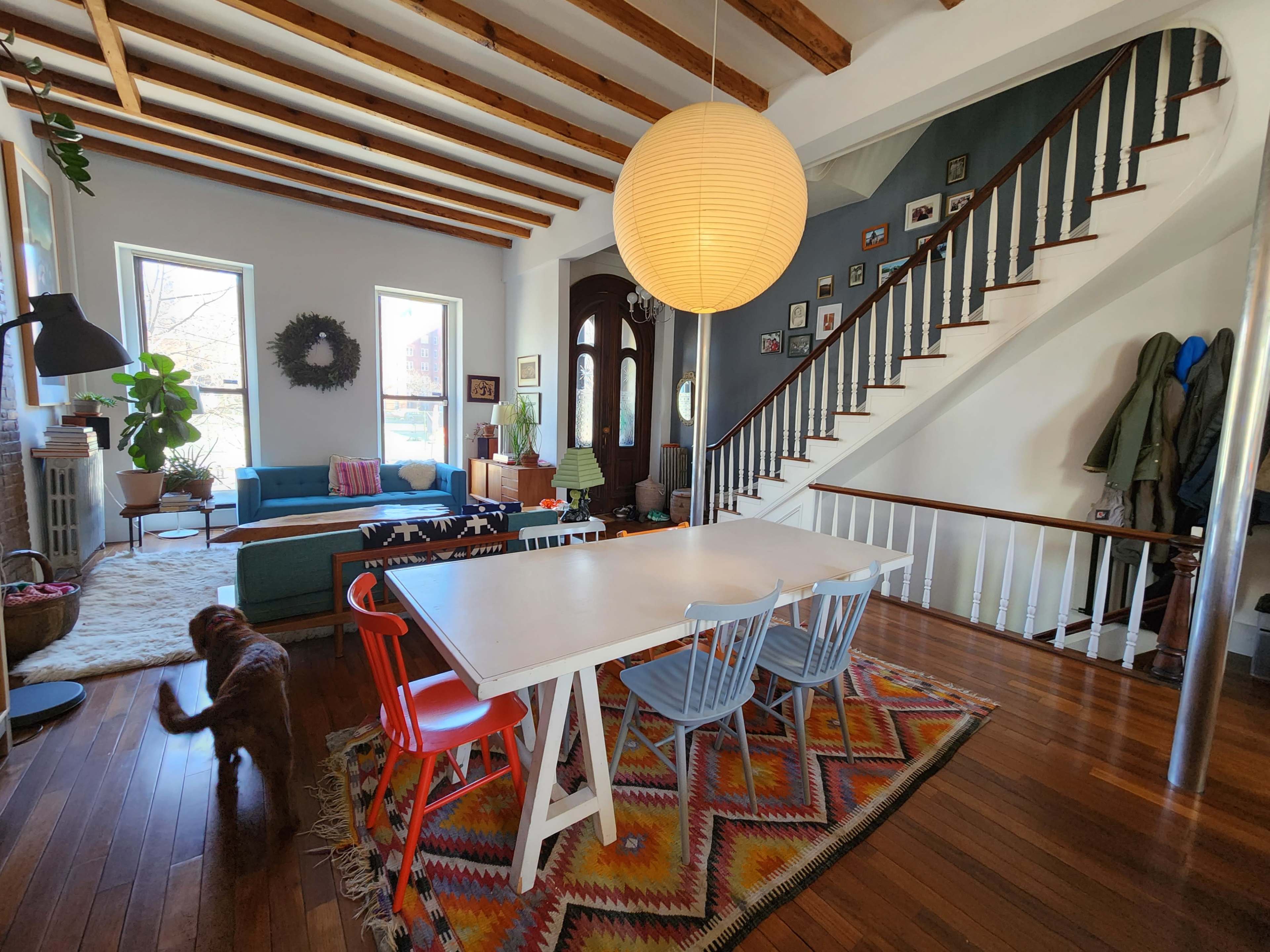 A spacious living and dining area features wooden beams, a large round pendant light, a colorful rug, and a dog in motion, alongside furniture arranged in a cozy layout.