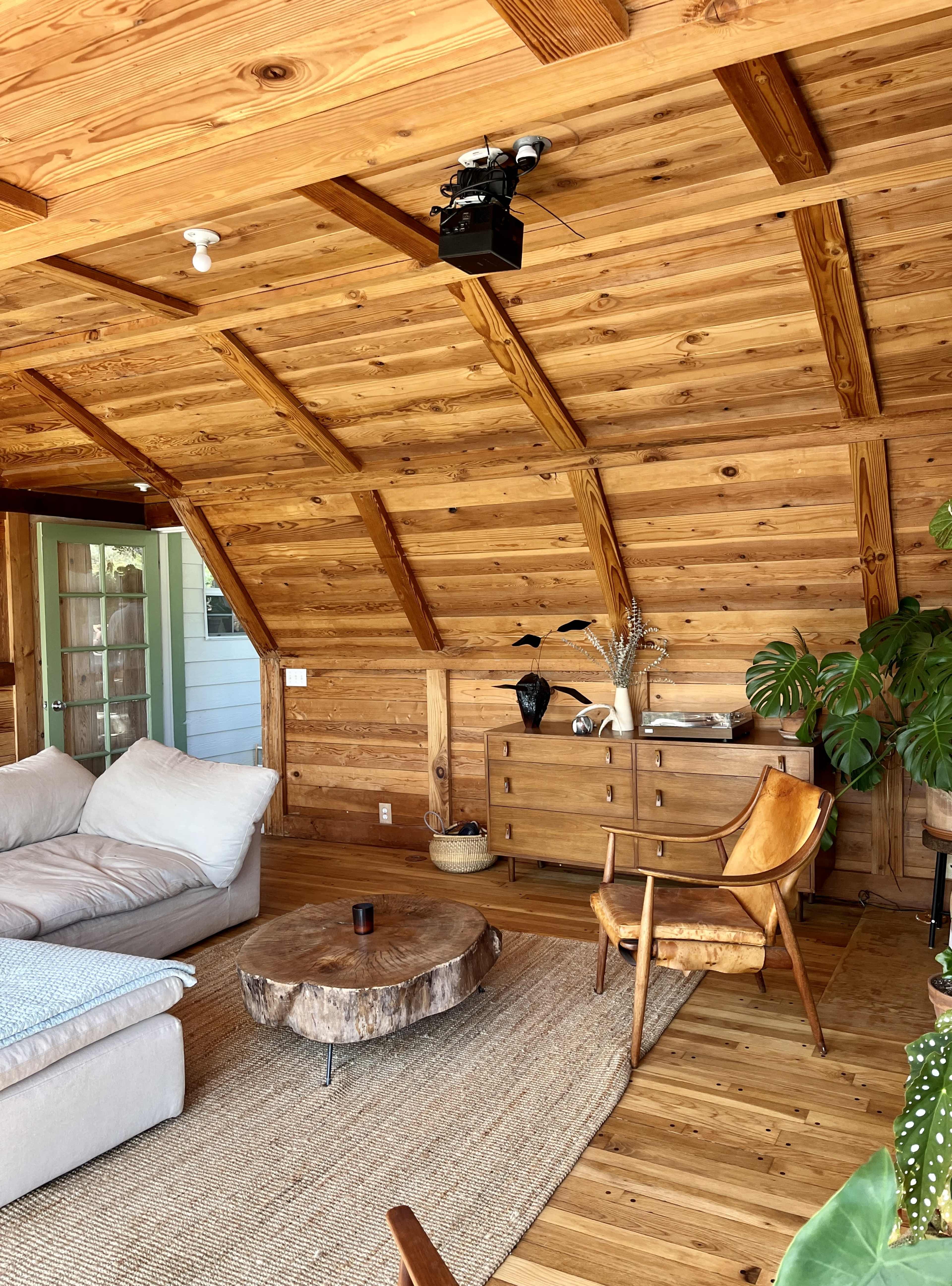 A cozy wooden interior with a sofa, a round coffee table made of tree trunk, a mid-century modern chair, and a cabinet against a wood-paneled wall.