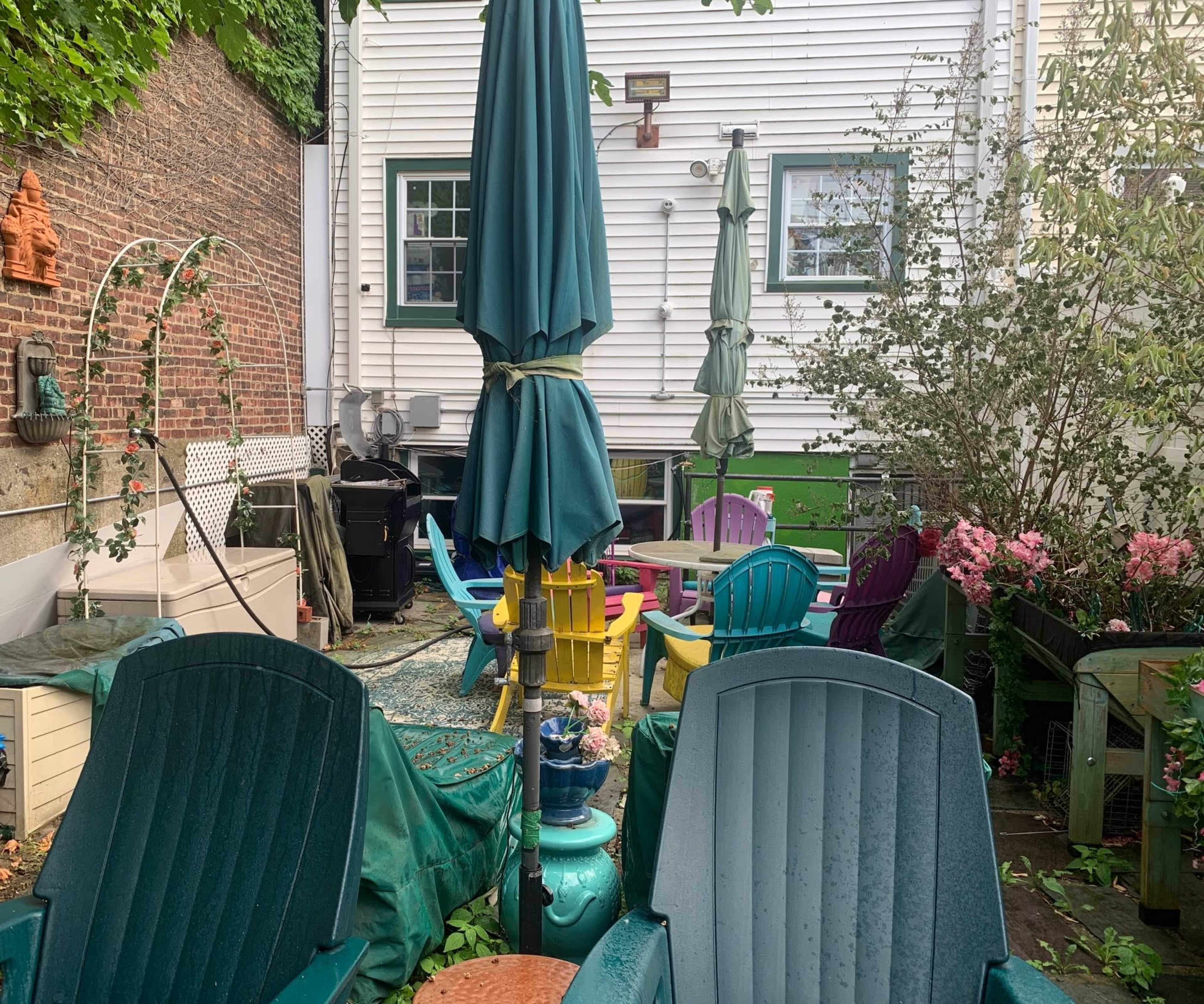 A backyard patio features various colorful chairs, tables, and umbrellas surrounded by greenery and a brick wall.
