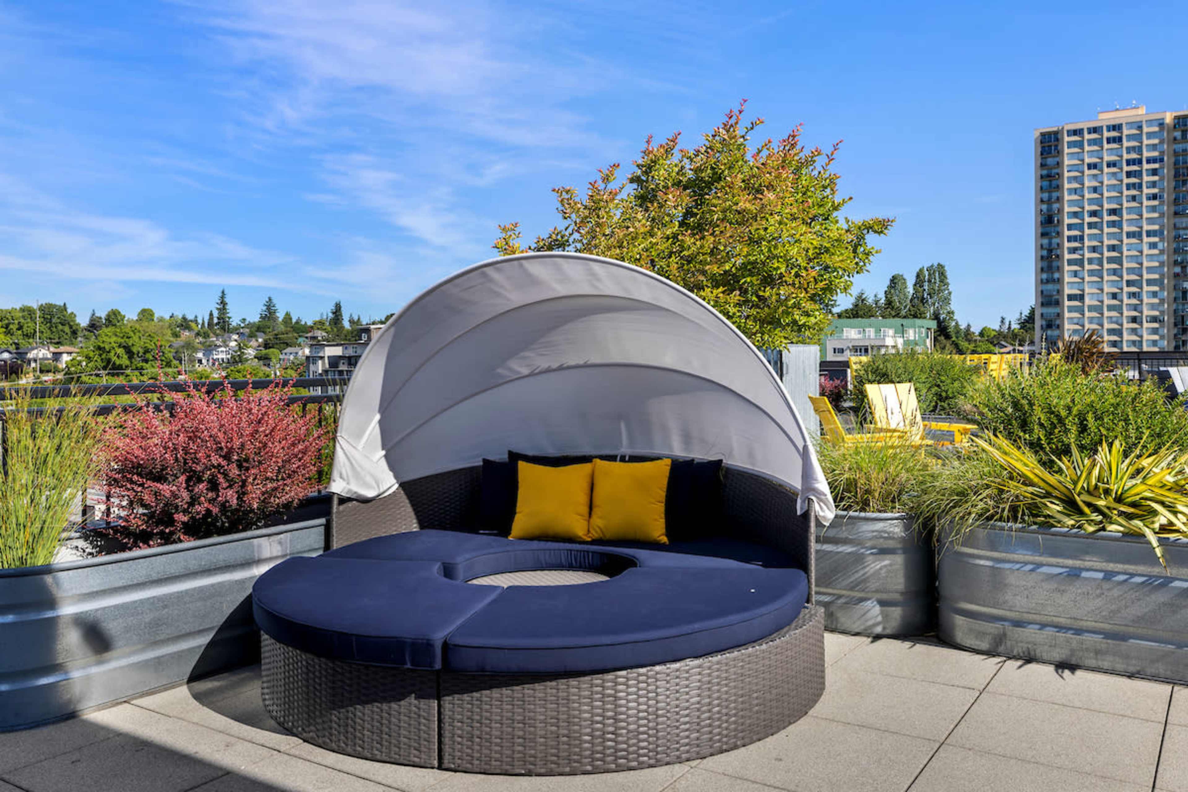 A circular outdoor lounge chair with a canopy is positioned on a paved deck surrounded by green vegetation and a tall building in the background.