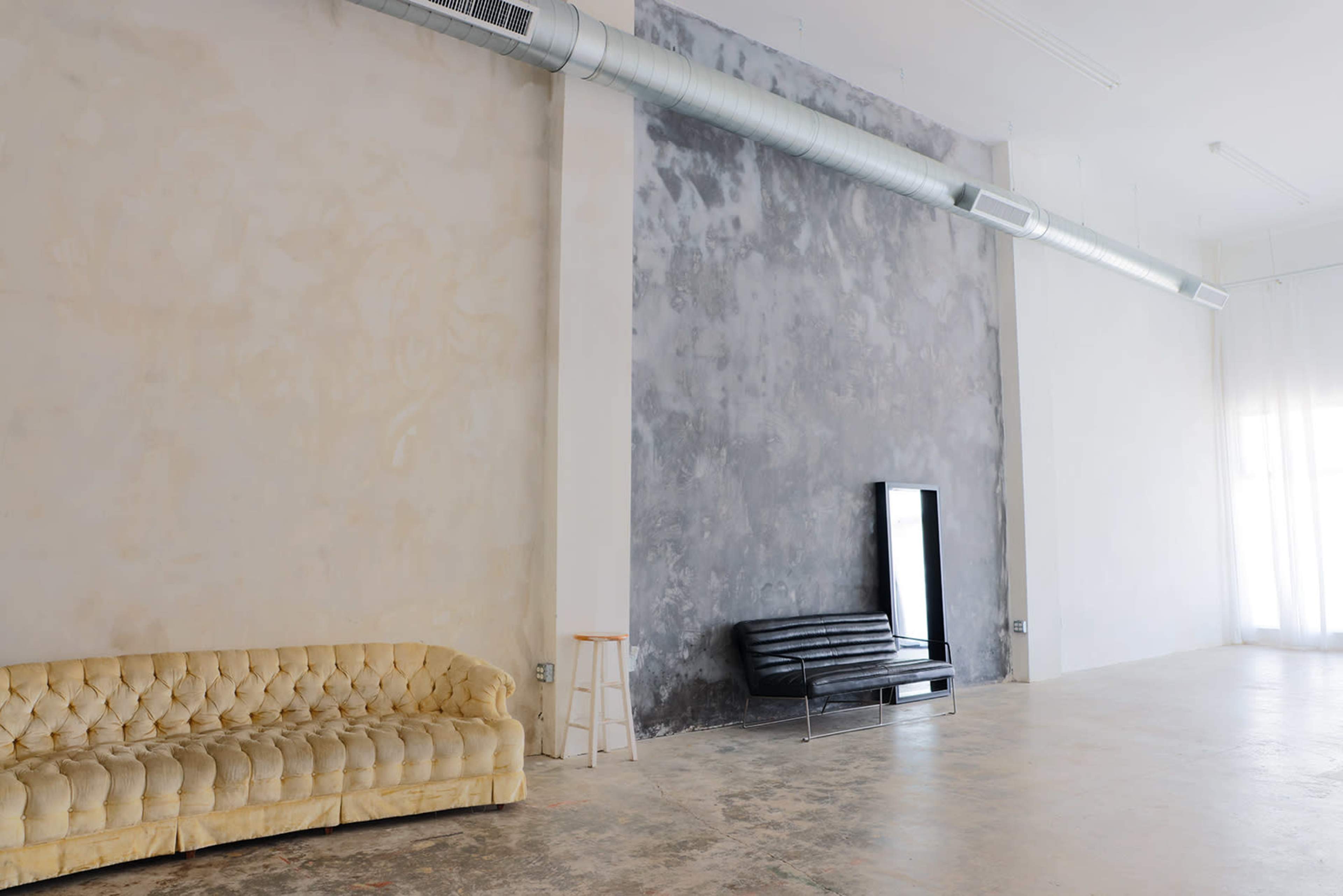 The image shows a spacious, minimalist room with a two-tone wall, a light-colored tufted sofa, a black leather couch, and a tall mirror against one wall.