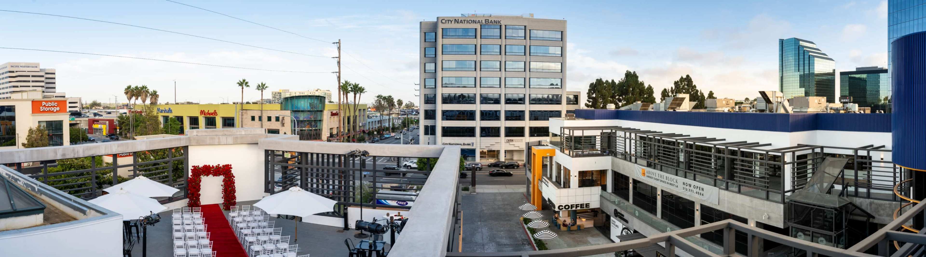 The image shows a panoramic view of a modern urban landscape with a bank building and various retail spaces along a street, along with outdoor seating and event setup on a rooftop terrace.