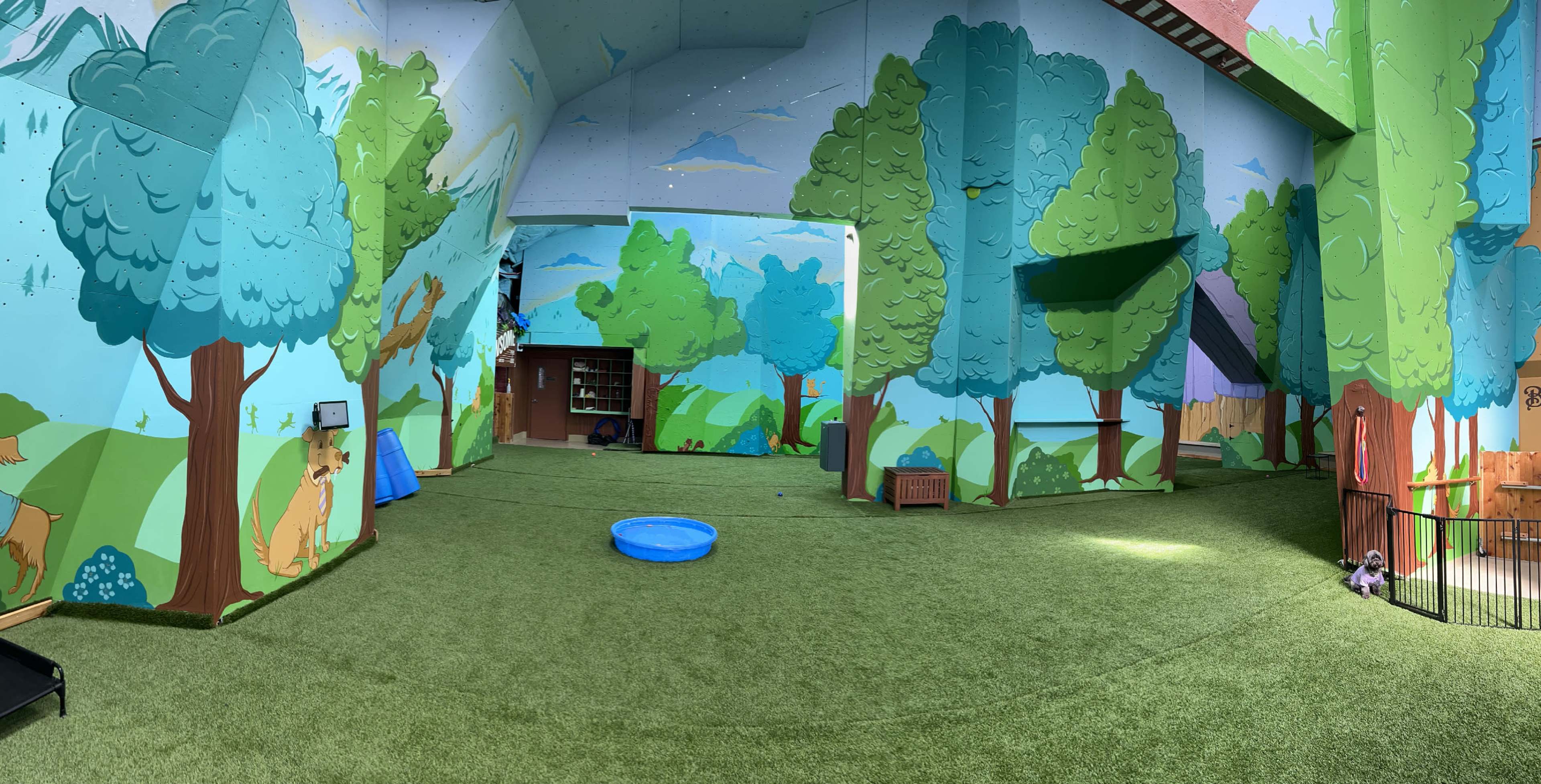The image shows a spacious indoor play area with artificial grass, adorned with vibrant murals of trees and nature.