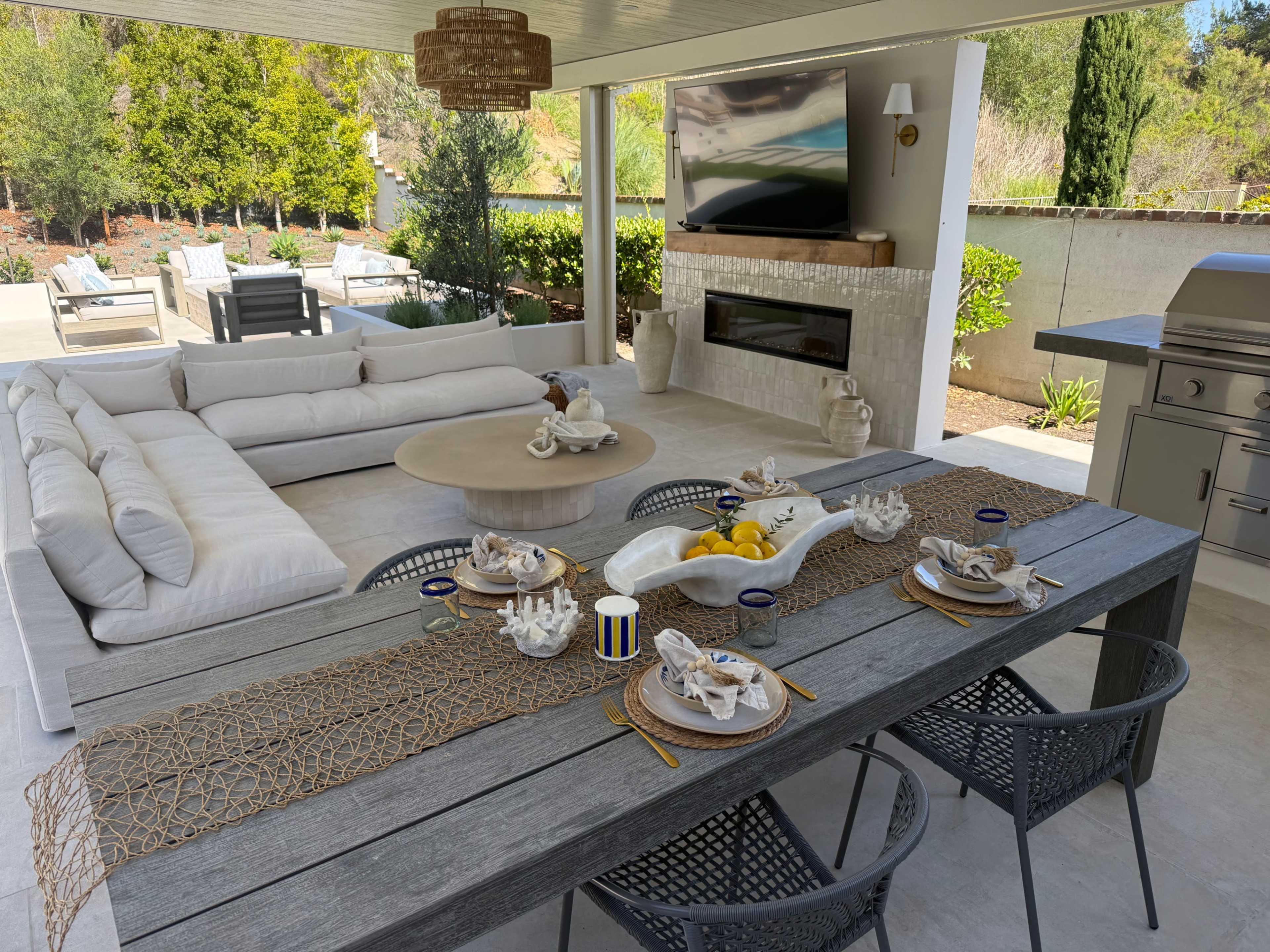 The image shows a stylish outdoor dining area featuring a wooden table set with dishware, a creamy sofa arrangement, and a large television mounted on the wall.