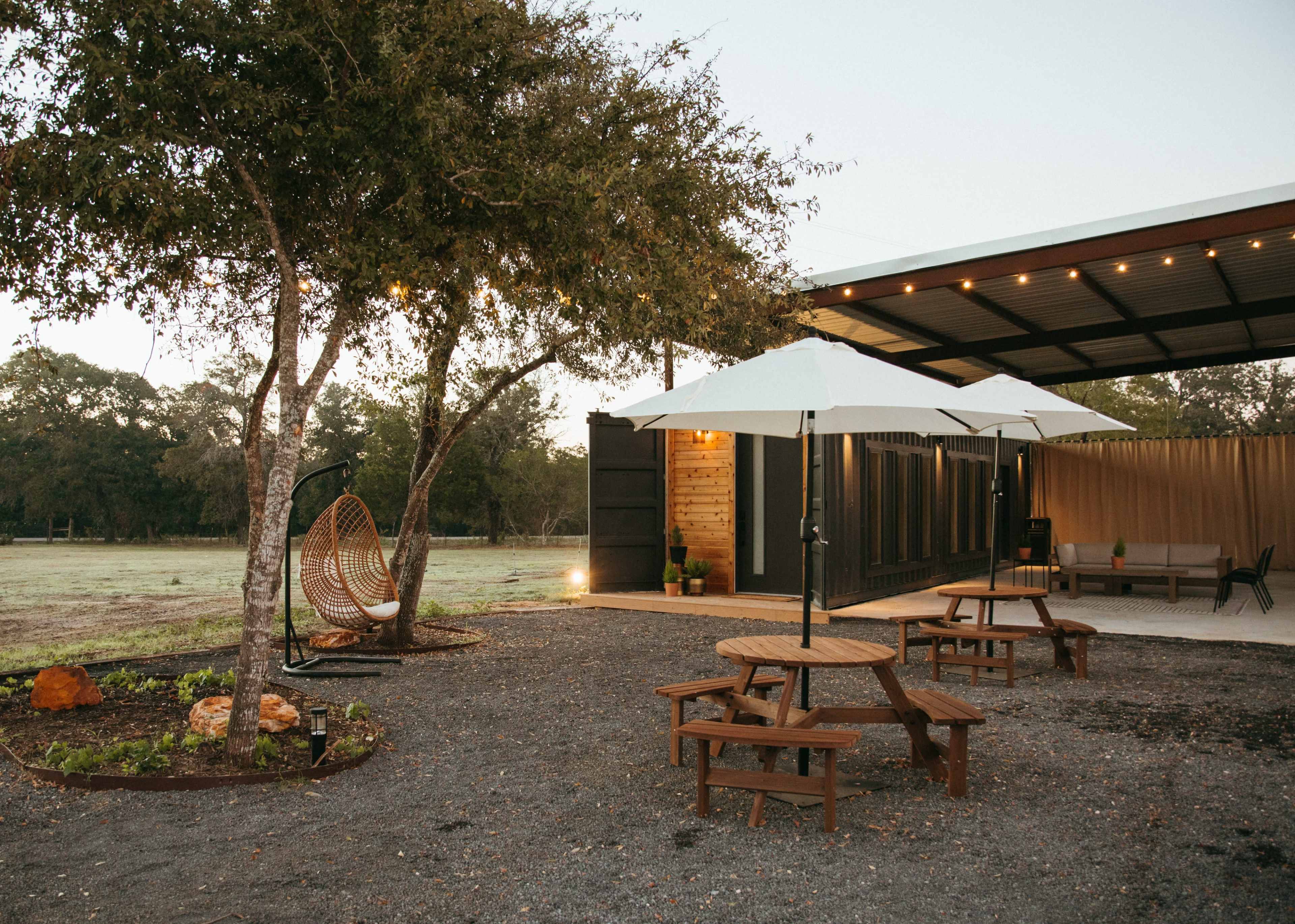 A modern outdoor space features a sleek, covered area with picnic tables, a hanging chair, and two large umbrellas, surrounded by gravel and landscaped plants.