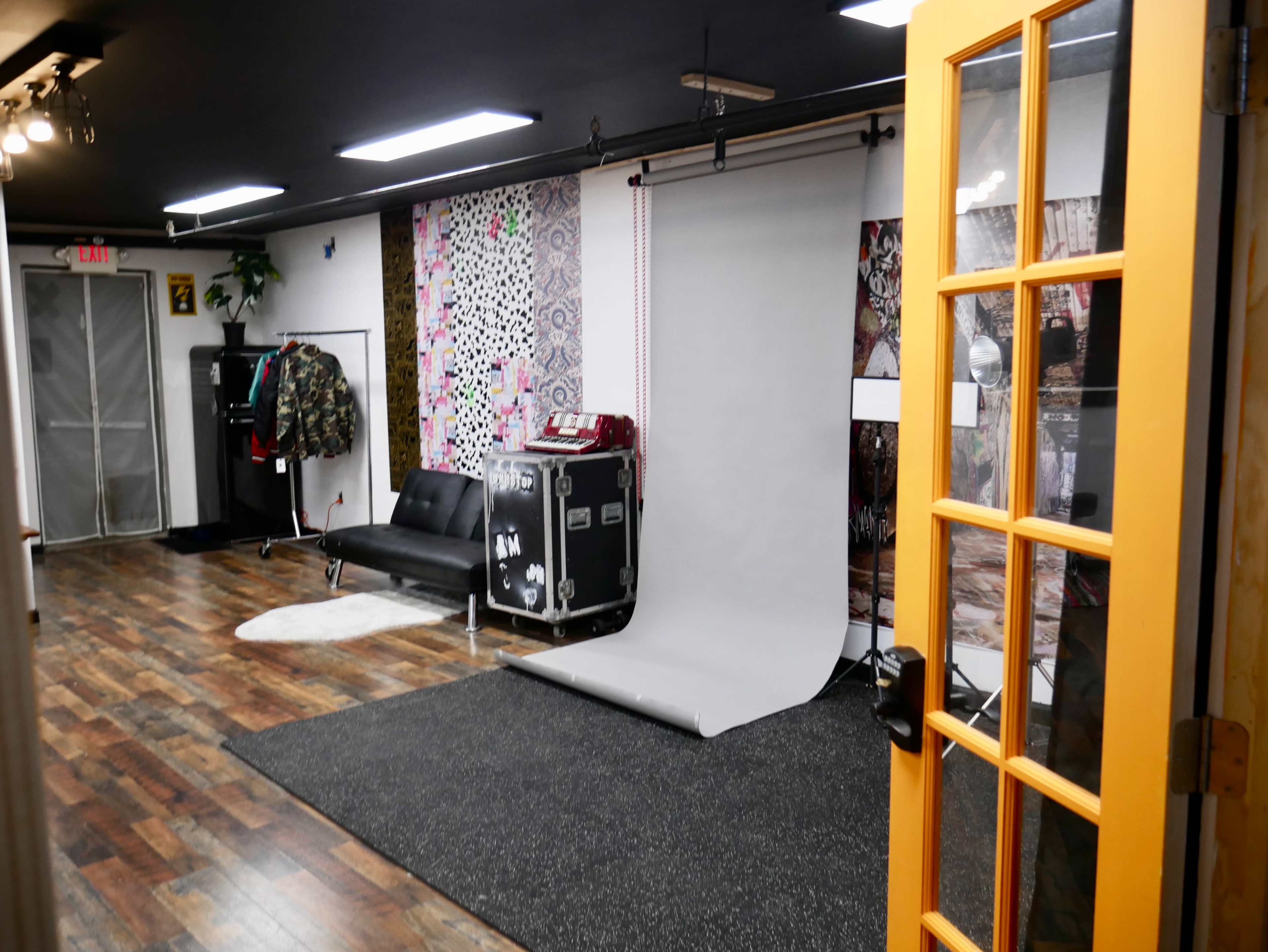 The image shows a stylish room with a gray backdrop, a black couch, colorful wallpaper, and equipment for a photoshoot.