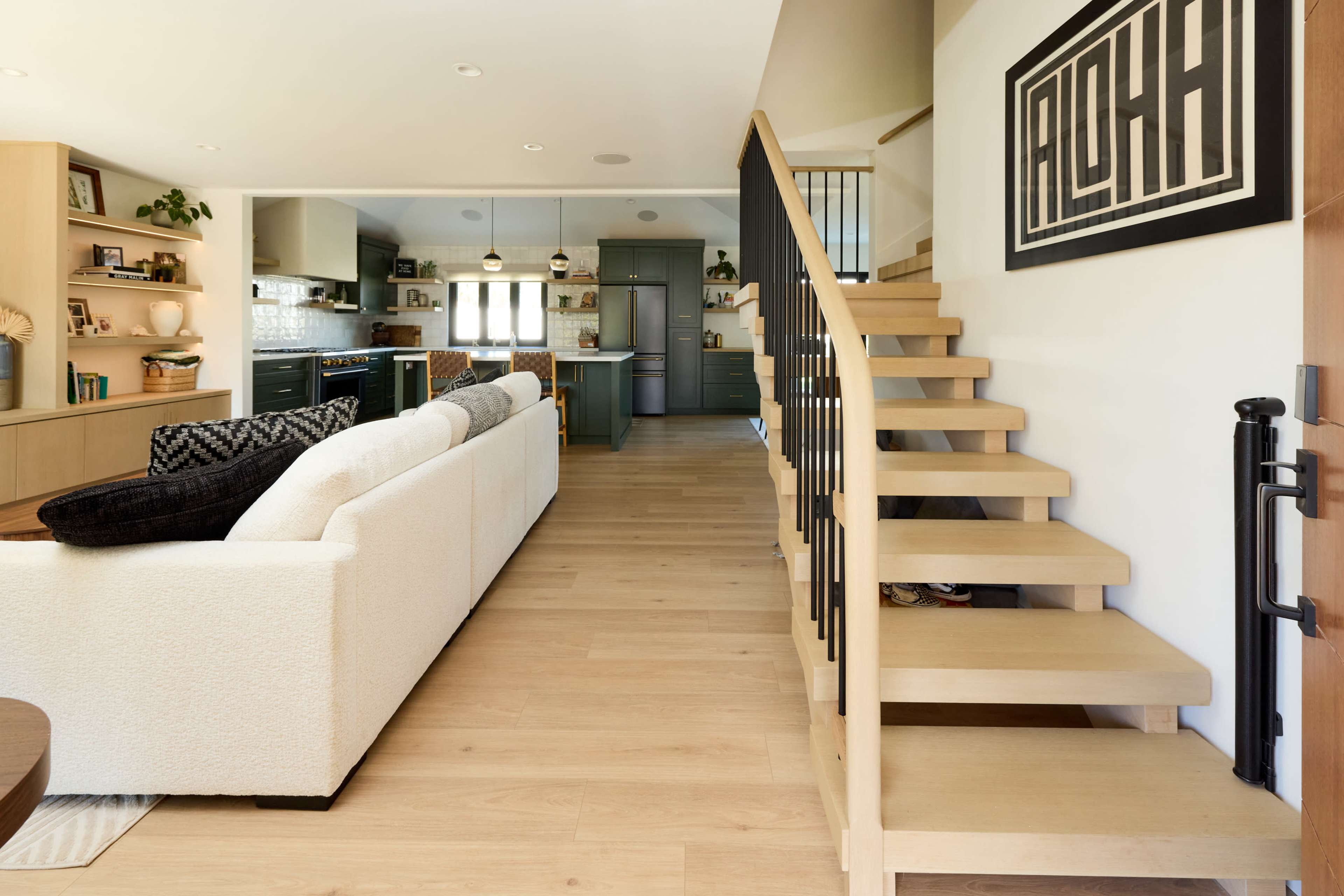 The image shows a modern interior of a house featuring a living area with a sofa, stairs leading to an upper floor, and a kitchen in the background.