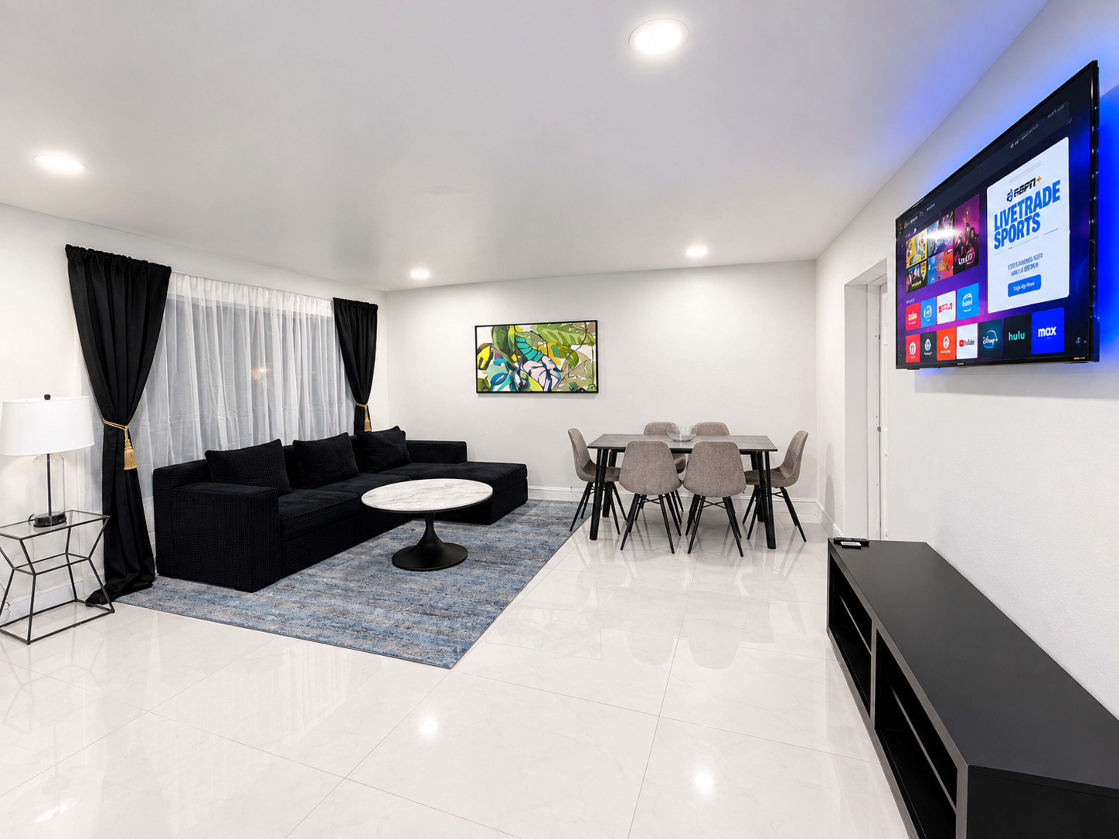 The image shows a modern living room with a dark-colored sectional sofa, a round marble coffee table, a dining area with a table and chairs, and a wall-mounted TV.