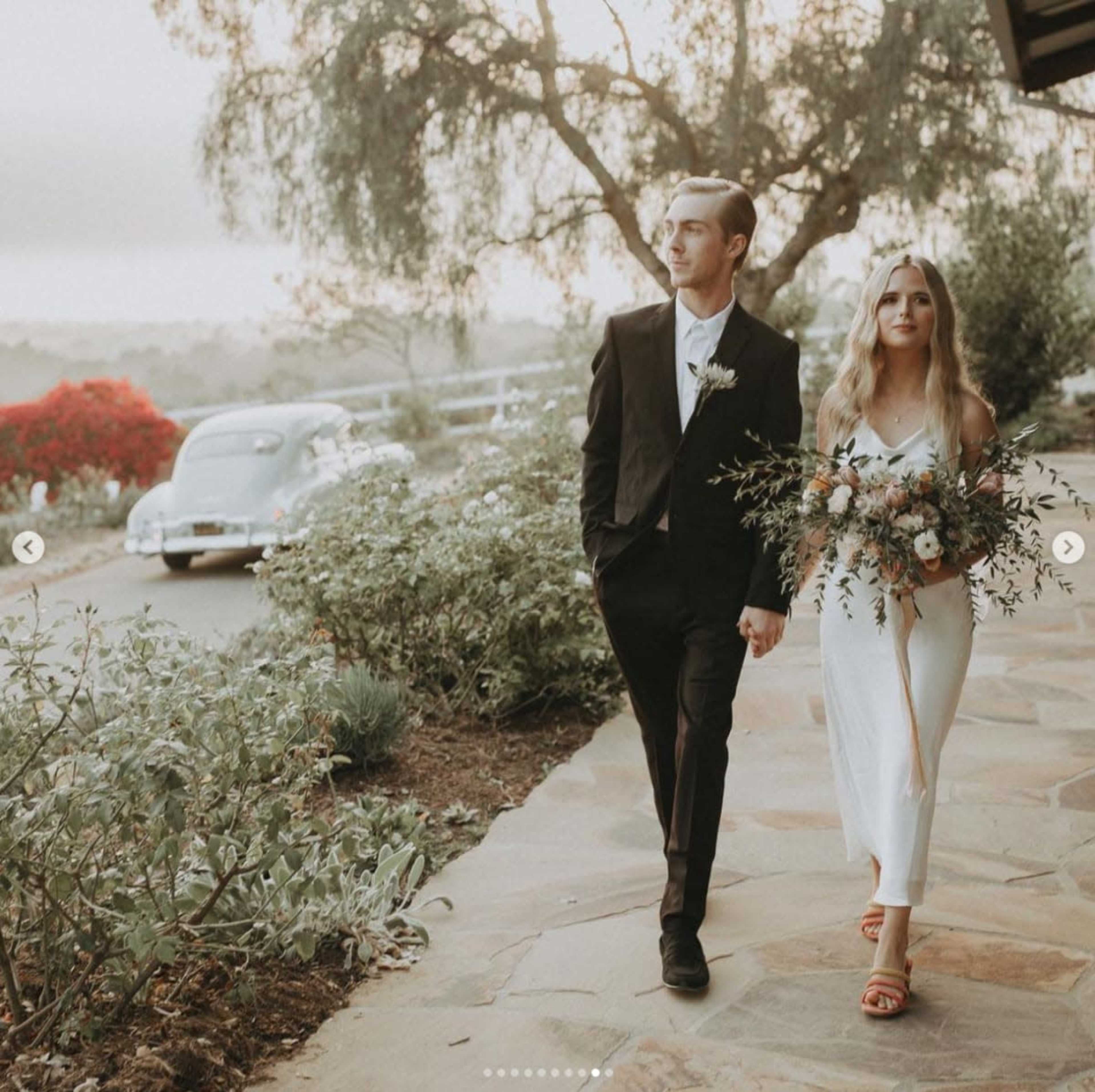 A couple walks hand in hand along a stone pathway lined with greenery, with a vintage car parked nearby and flowering bushes in the background.