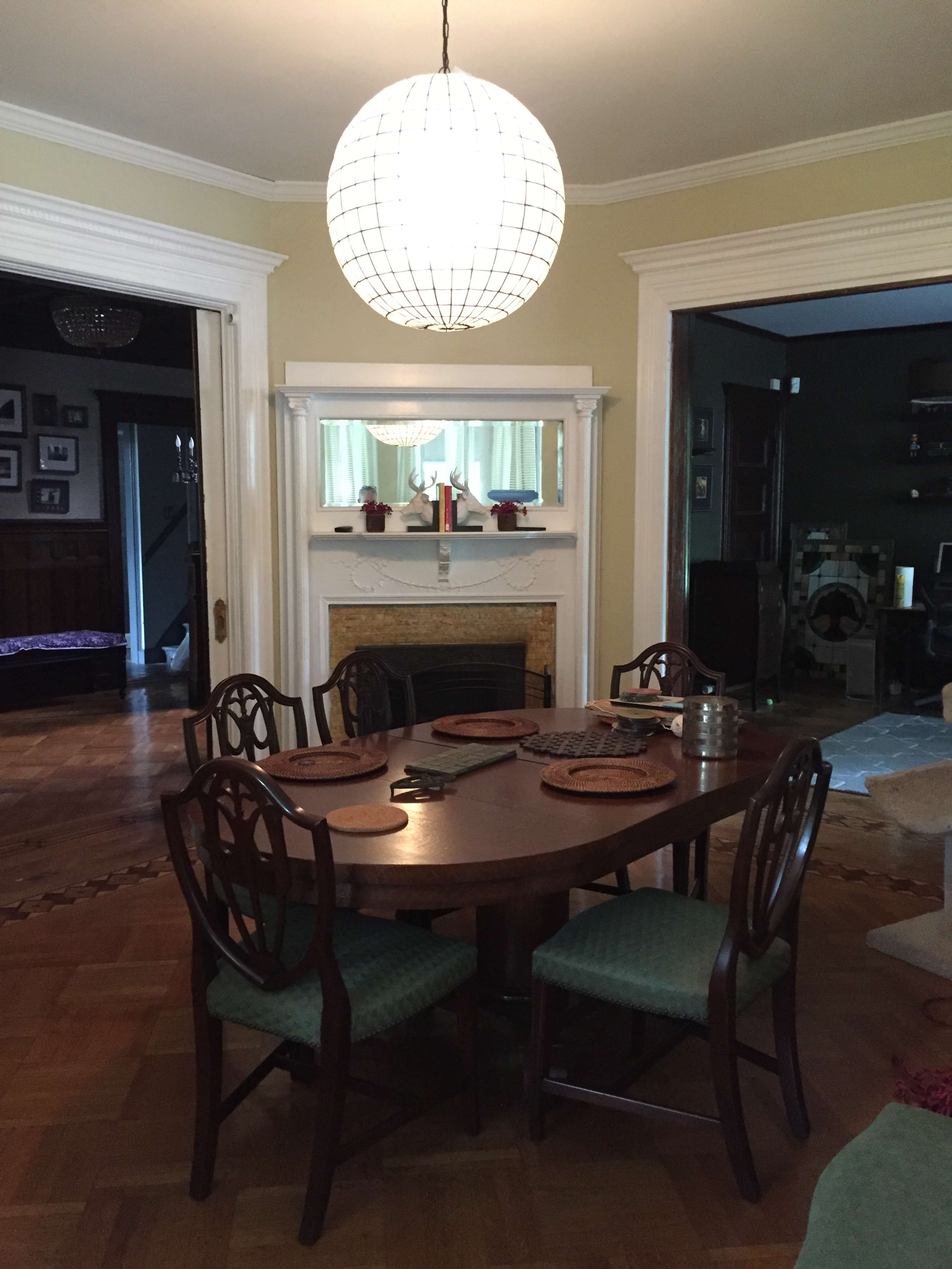 A round wooden dining table is set in a well-lit room, surrounded by four chairs, beneath a large pendant light.