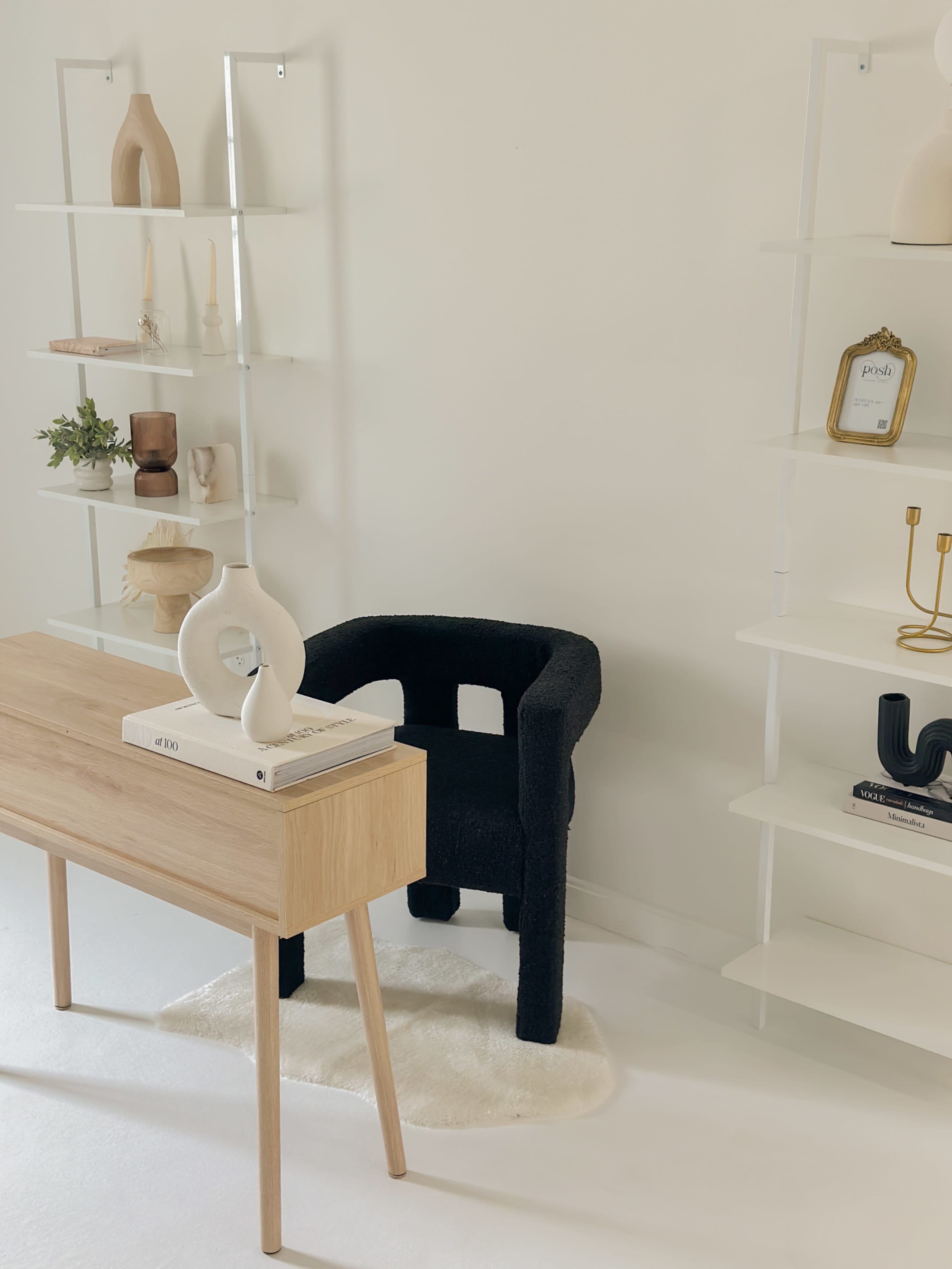 A minimalist workspace features a wooden desk with books and a decorative object, a black upholstered chair, and white shelving displaying various decor items.