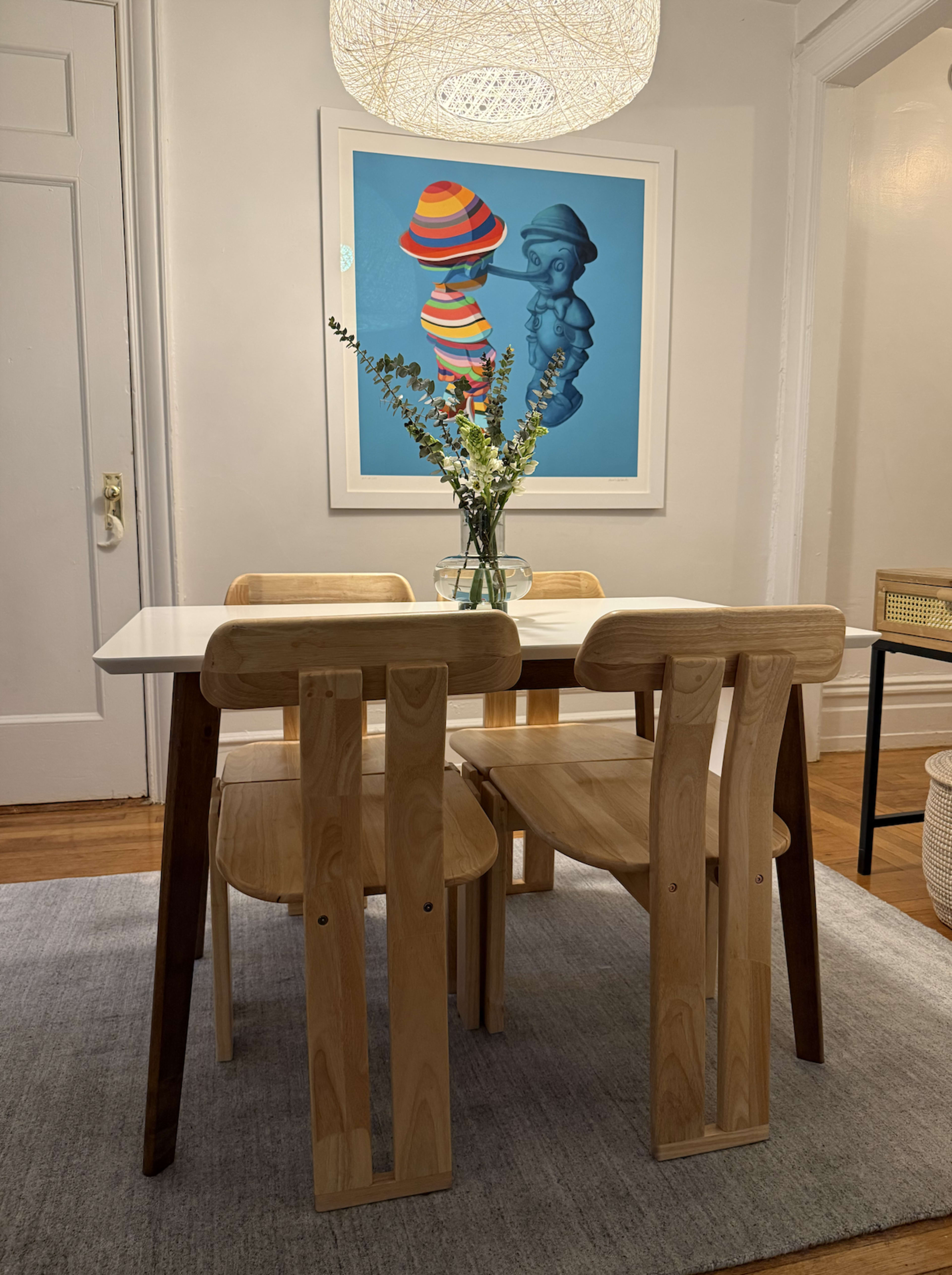 A modern dining table with four wooden chairs is placed on a gray rug in a well-lit room featuring framed artwork on the wall.