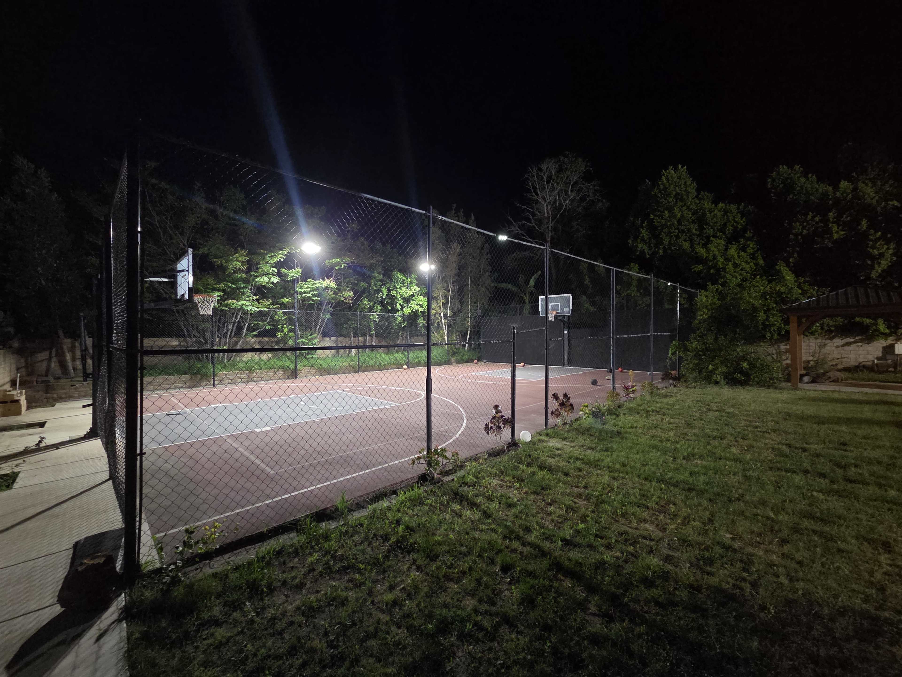 The image shows a well-lit outdoor basketball court enclosed by a chain-link fence, surrounded by trees in a nighttime setting.