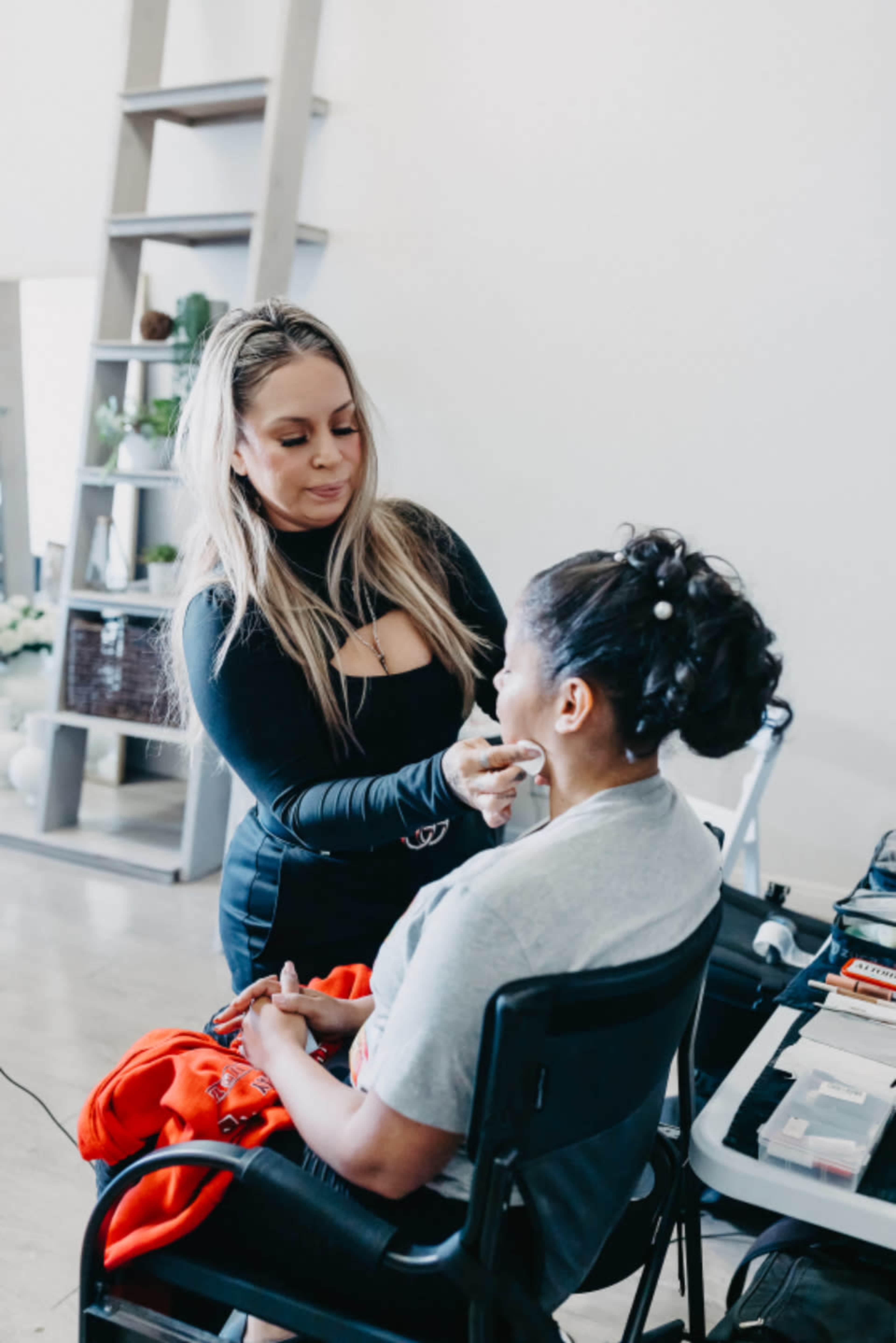 A makeup artist applies makeup to a seated client in a well-lit room with a modern aesthetic.