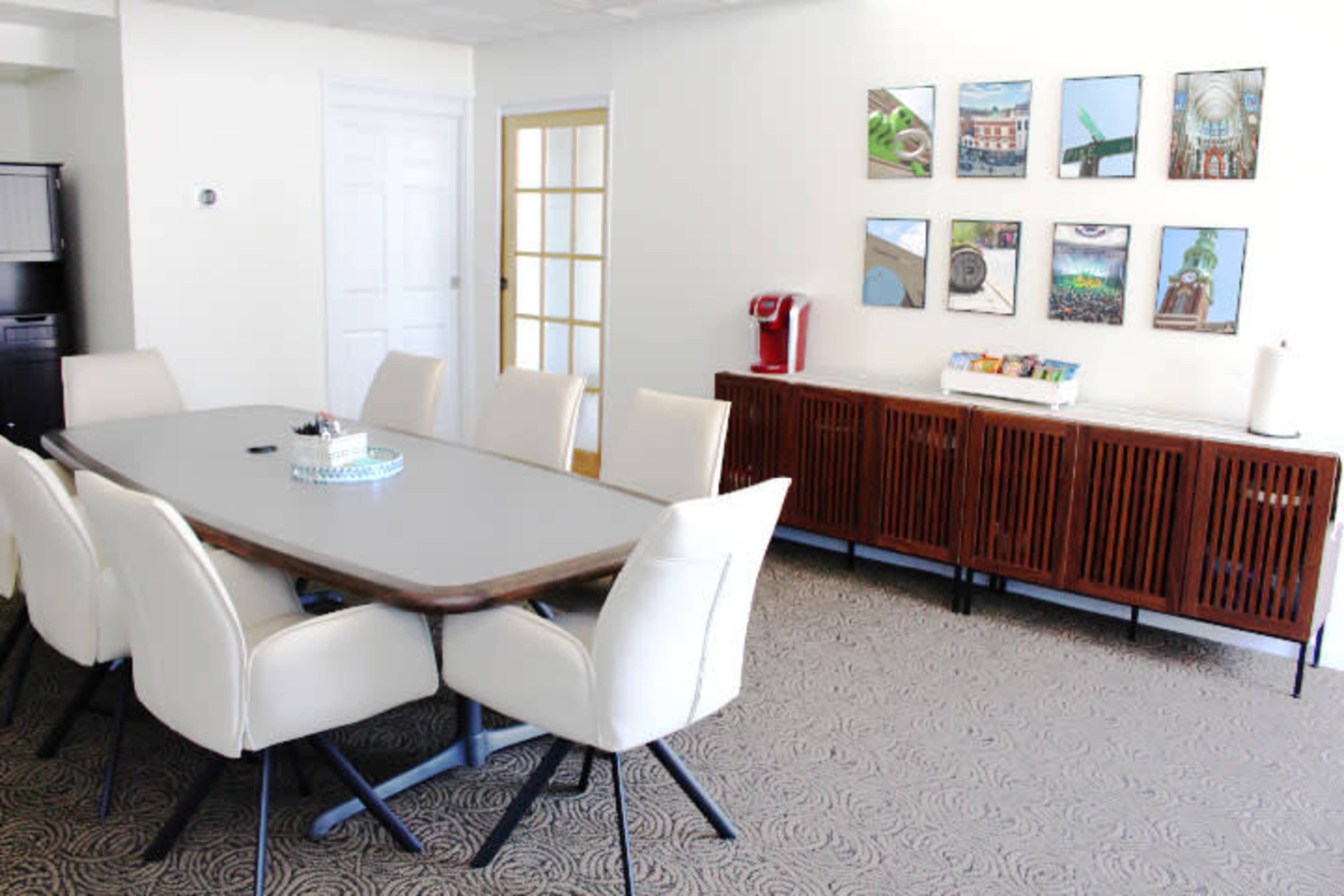 A modern conference room features a long table surrounded by beige chairs, with a side cabinet displaying art on the wall.