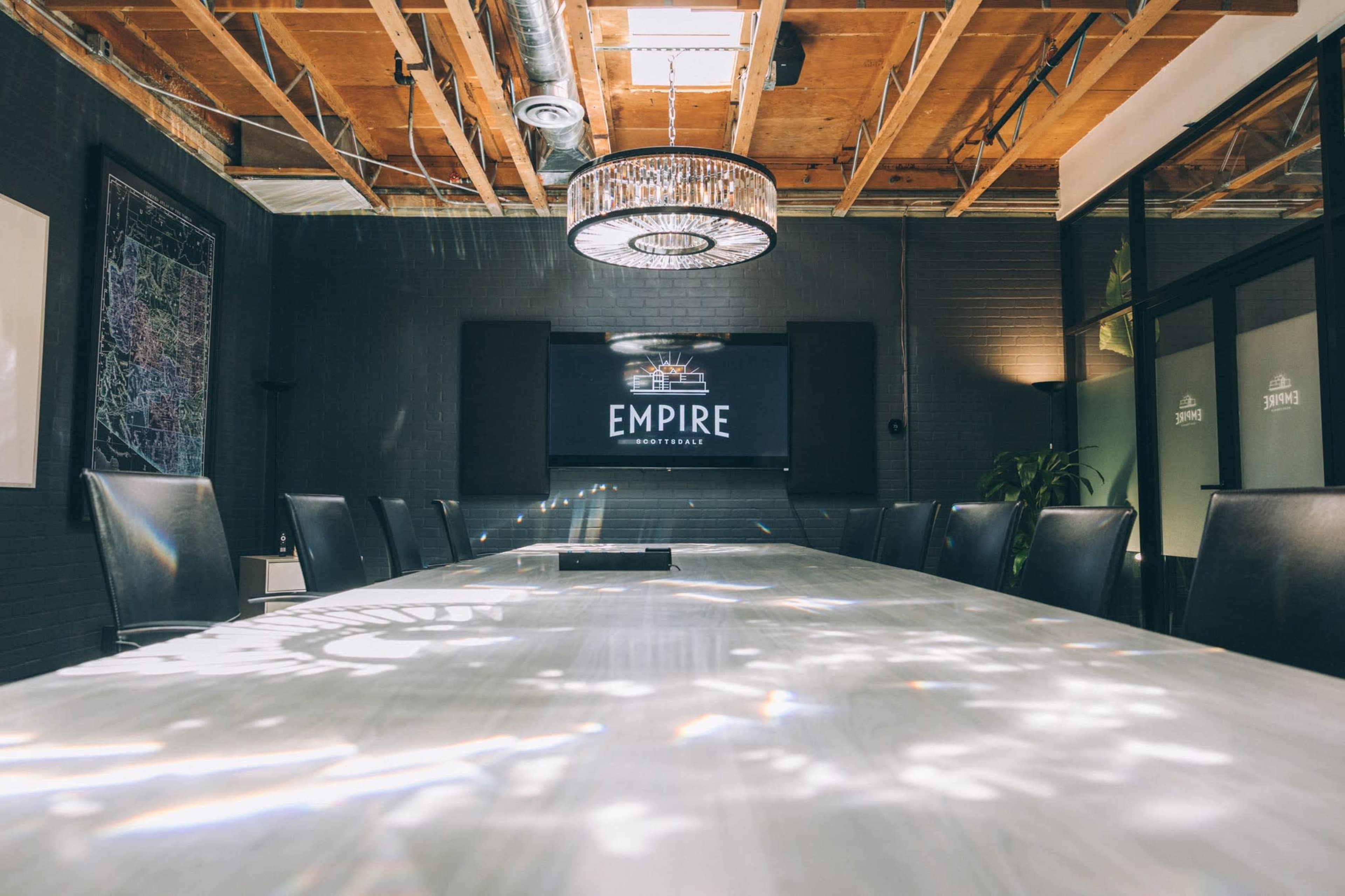 A modern conference room features a long table, black leather chairs, and a large screen displaying the word "EMPIRE" under a decorative light fixture.