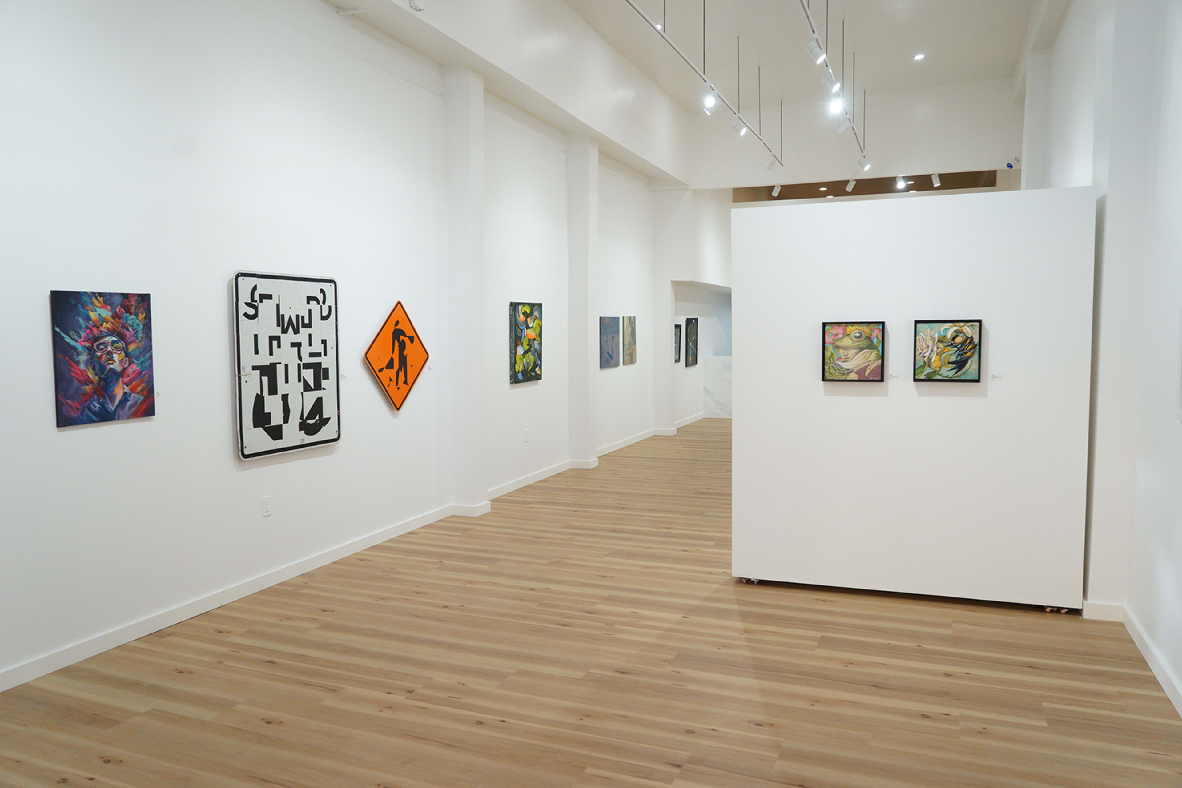 The image shows an art gallery interior with various colorful paintings displayed on the walls and a central white partition featuring additional artwork.