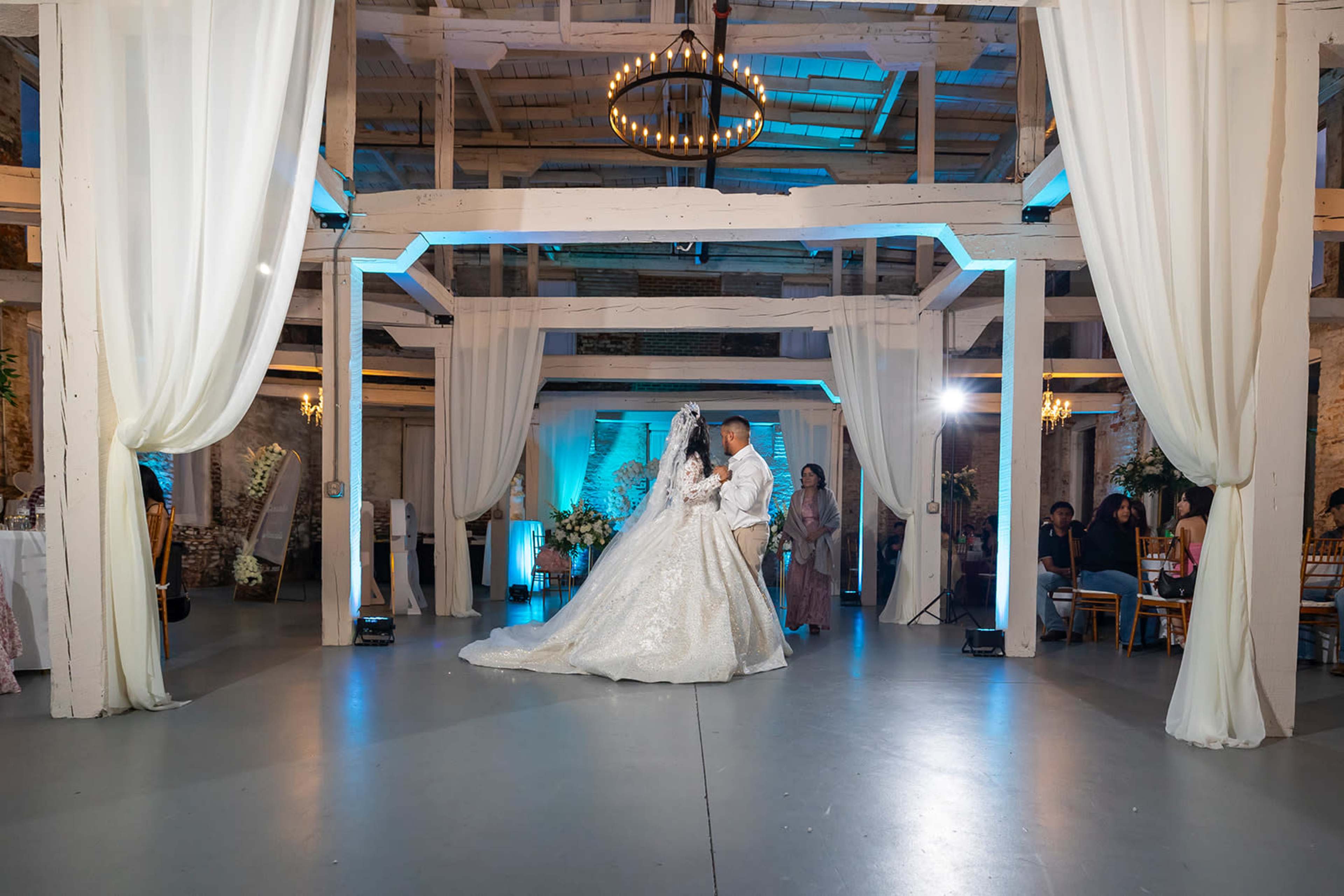 A bride and her partner share a dance in a decorated barn venue with hanging lights and draped fabric.