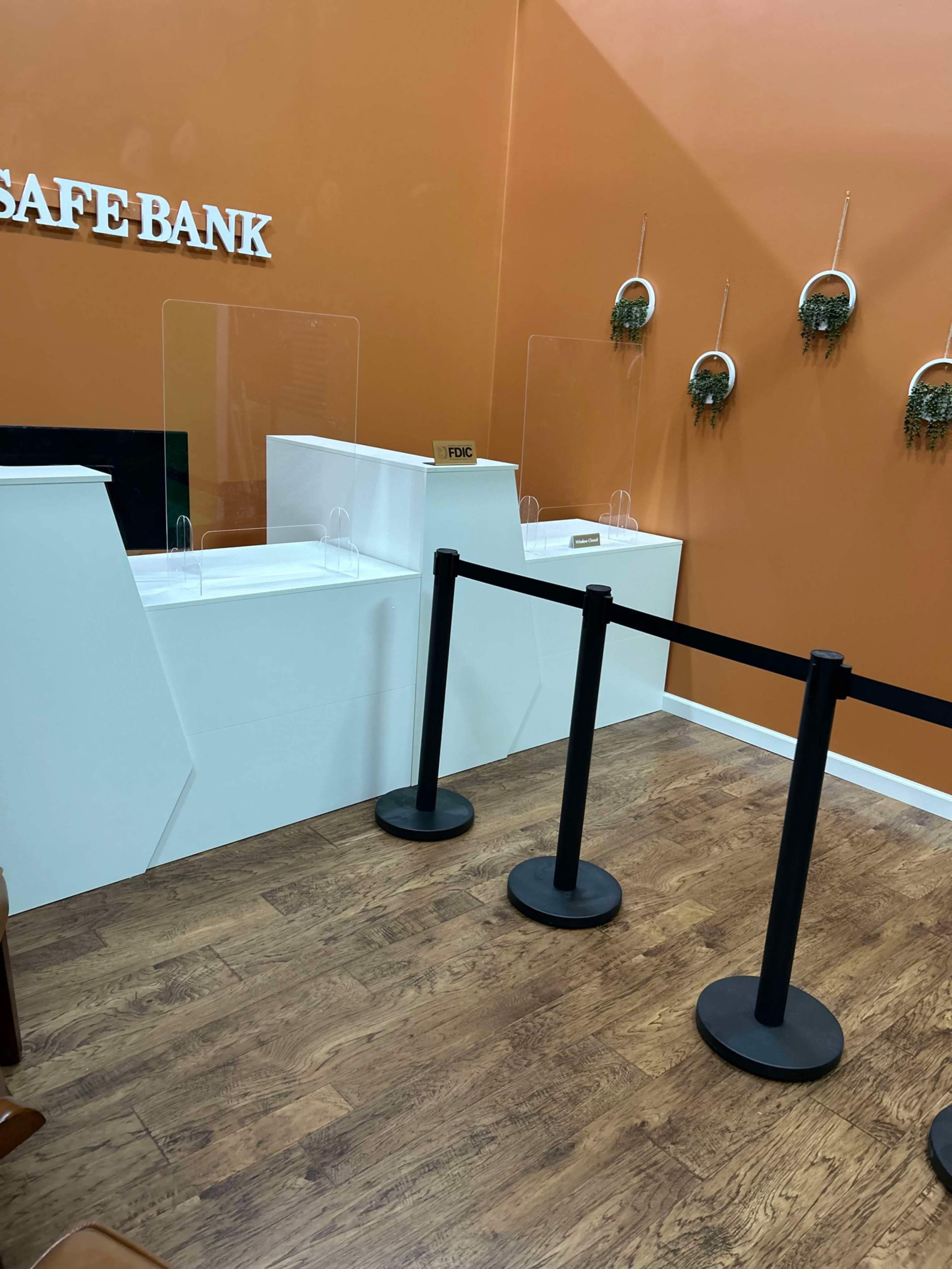The image shows a bank interior featuring a service counter with clear acrylic barriers and a line of stanchions separating the customer area.