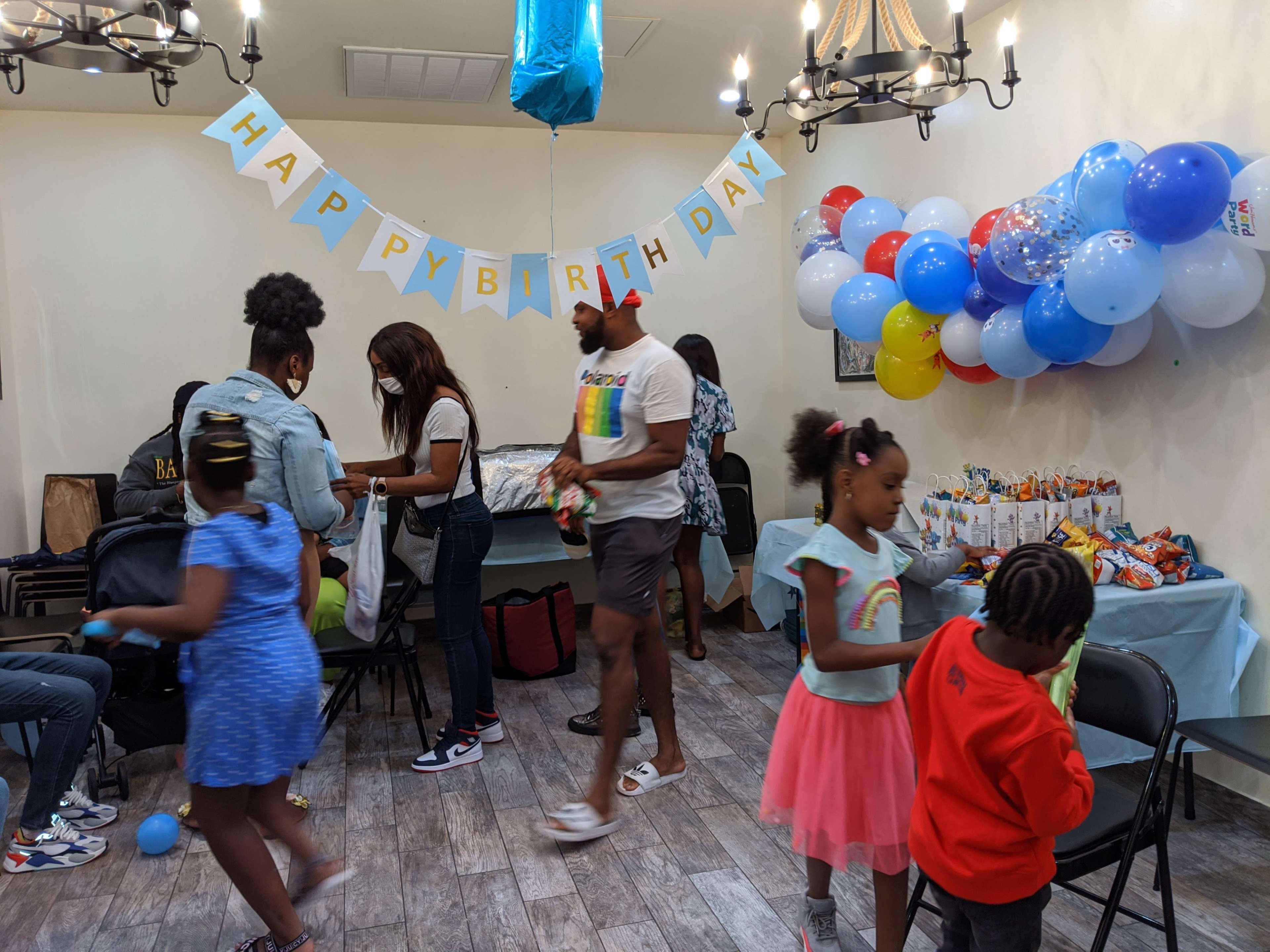 A group of people, including children, are gathered in a decorated room celebrating a birthday, with a balloon arch and a "Happy Birthday" banner.