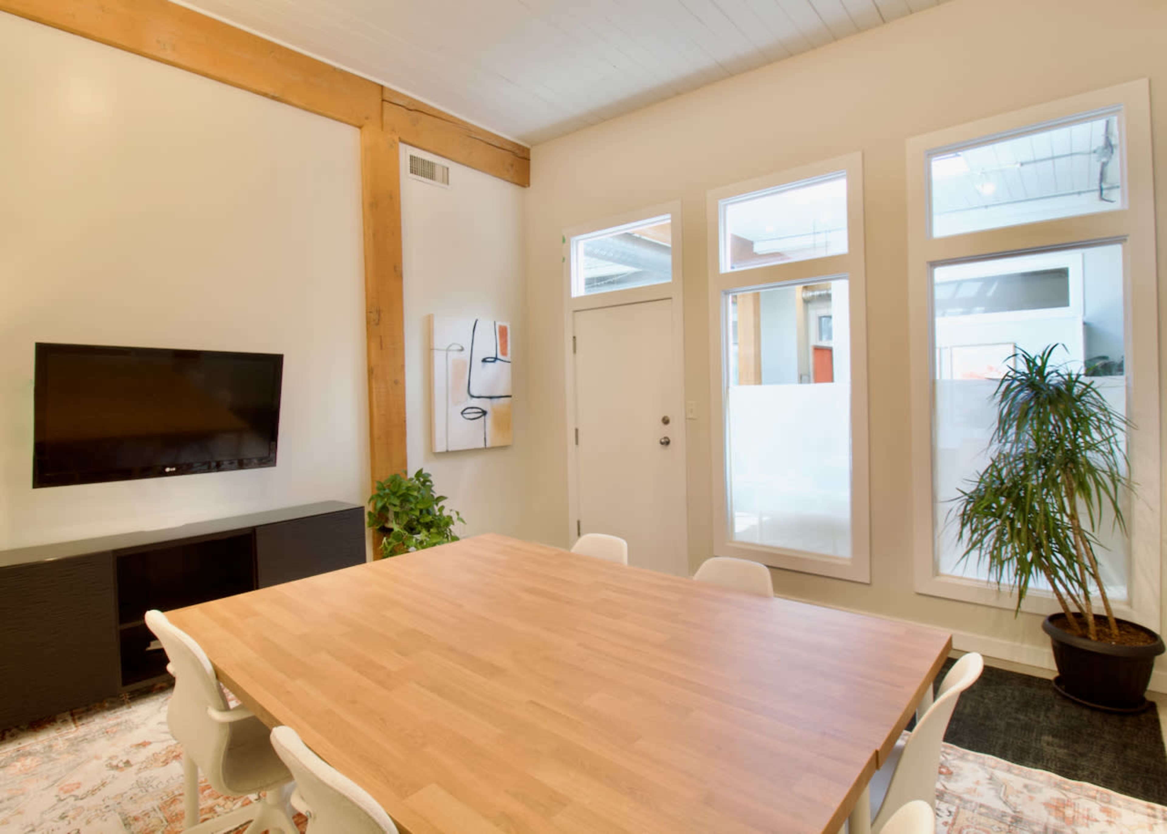 The image shows a modern meeting room with a large wooden table, white chairs, a wall-mounted TV, and large windows allowing natural light to enter.