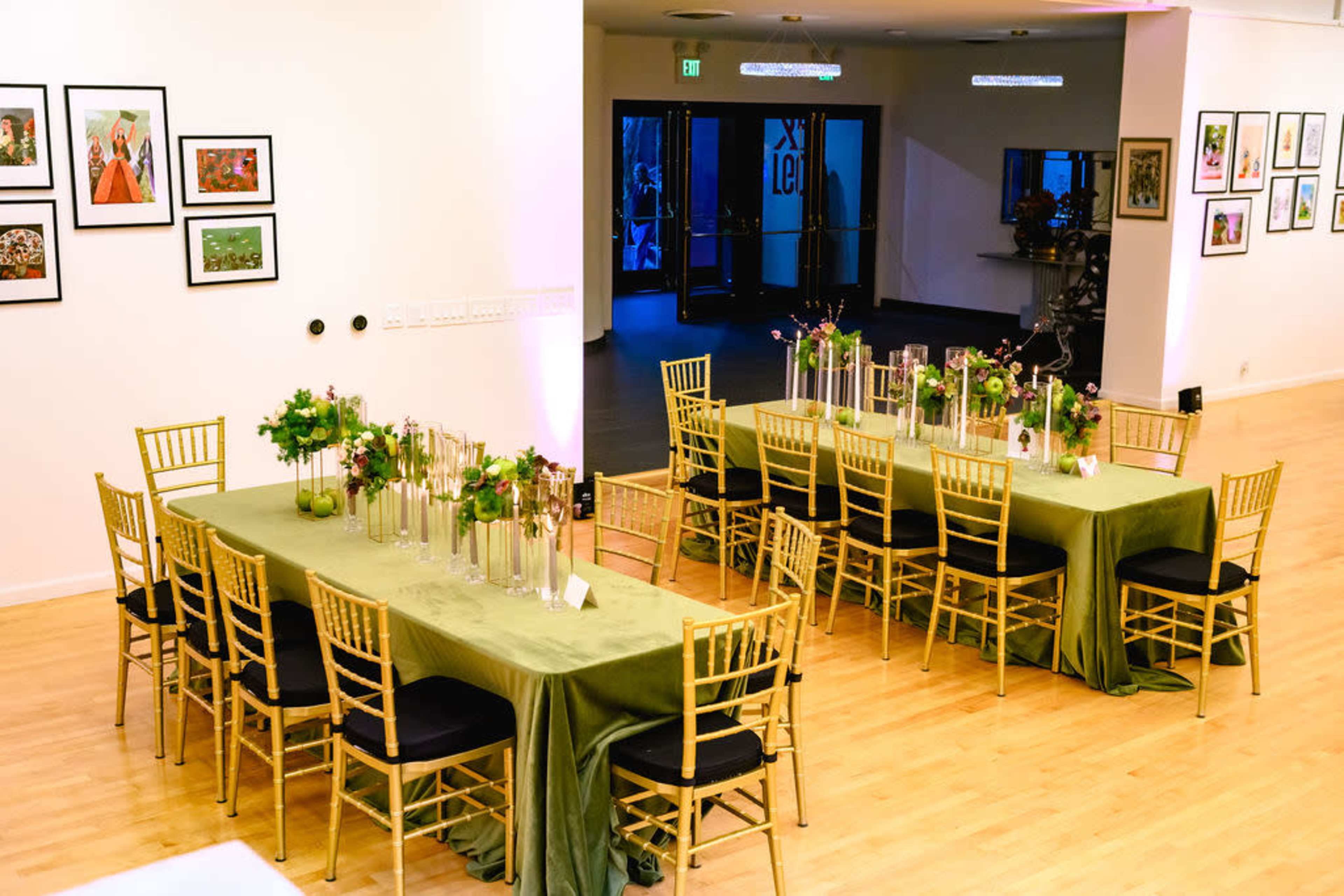 The image shows a spacious event room with two long tables set with green tablecloths, gold chairs, and floral centerpieces, facing a wall of framed artwork.