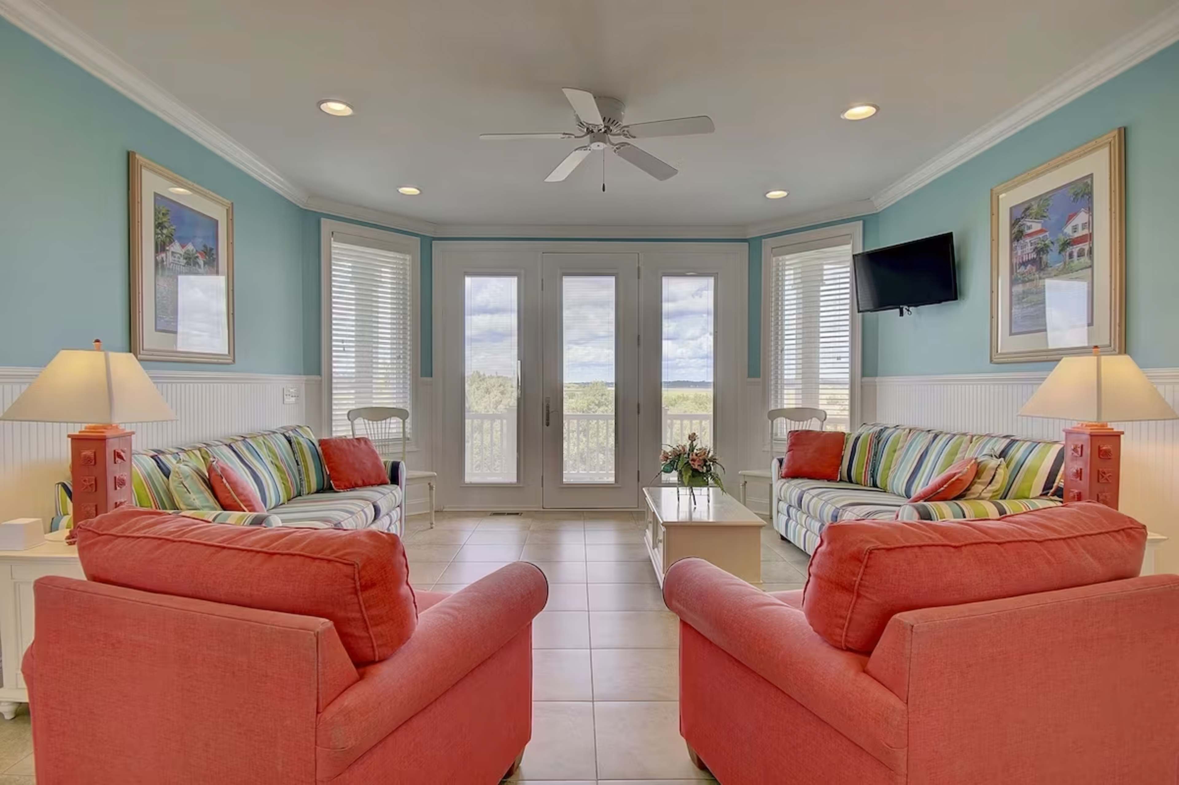 A bright and airy living room features two coral-colored armchairs, striped sofas, and large windows overlooking a scenic view.