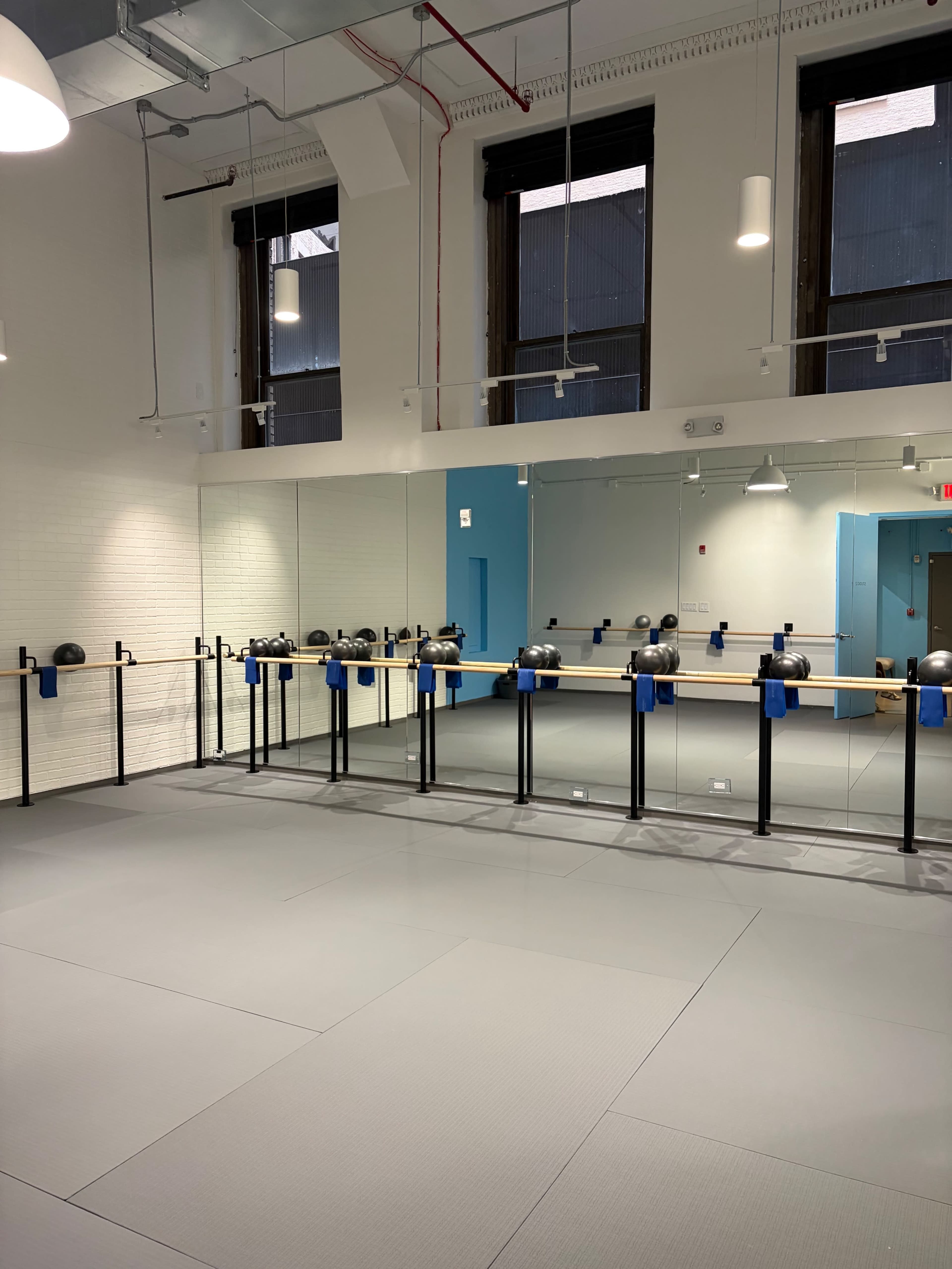 The image shows a spacious fitness studio featuring a wall of mirrors, a ballet barre, and a gray floor with blue accents.