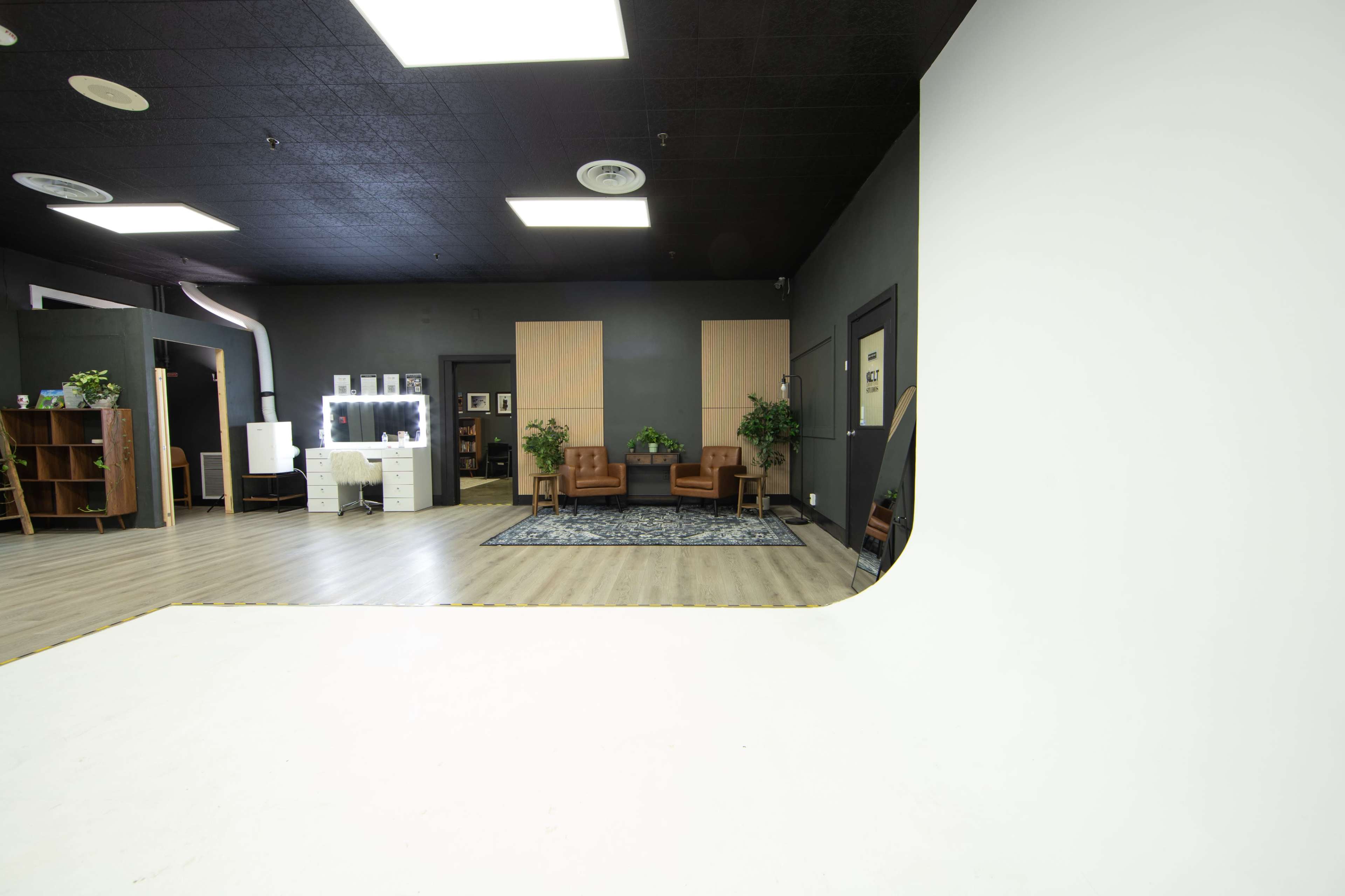The image shows a photography studio with a large white backdrop, two brown chairs, and plants arranged in a modern interior space.