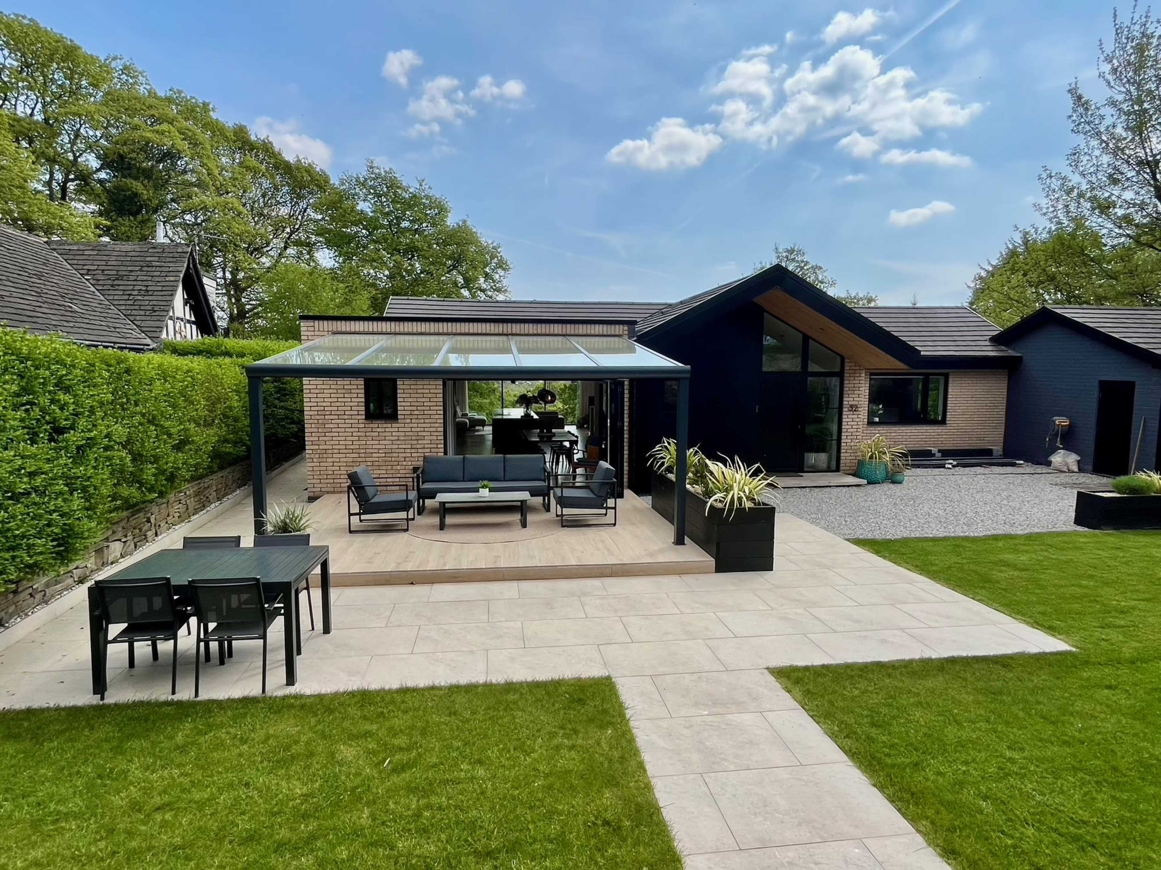 The image shows a modern house with a green lawn, a patio covered by a glass structure, and outdoor furniture.