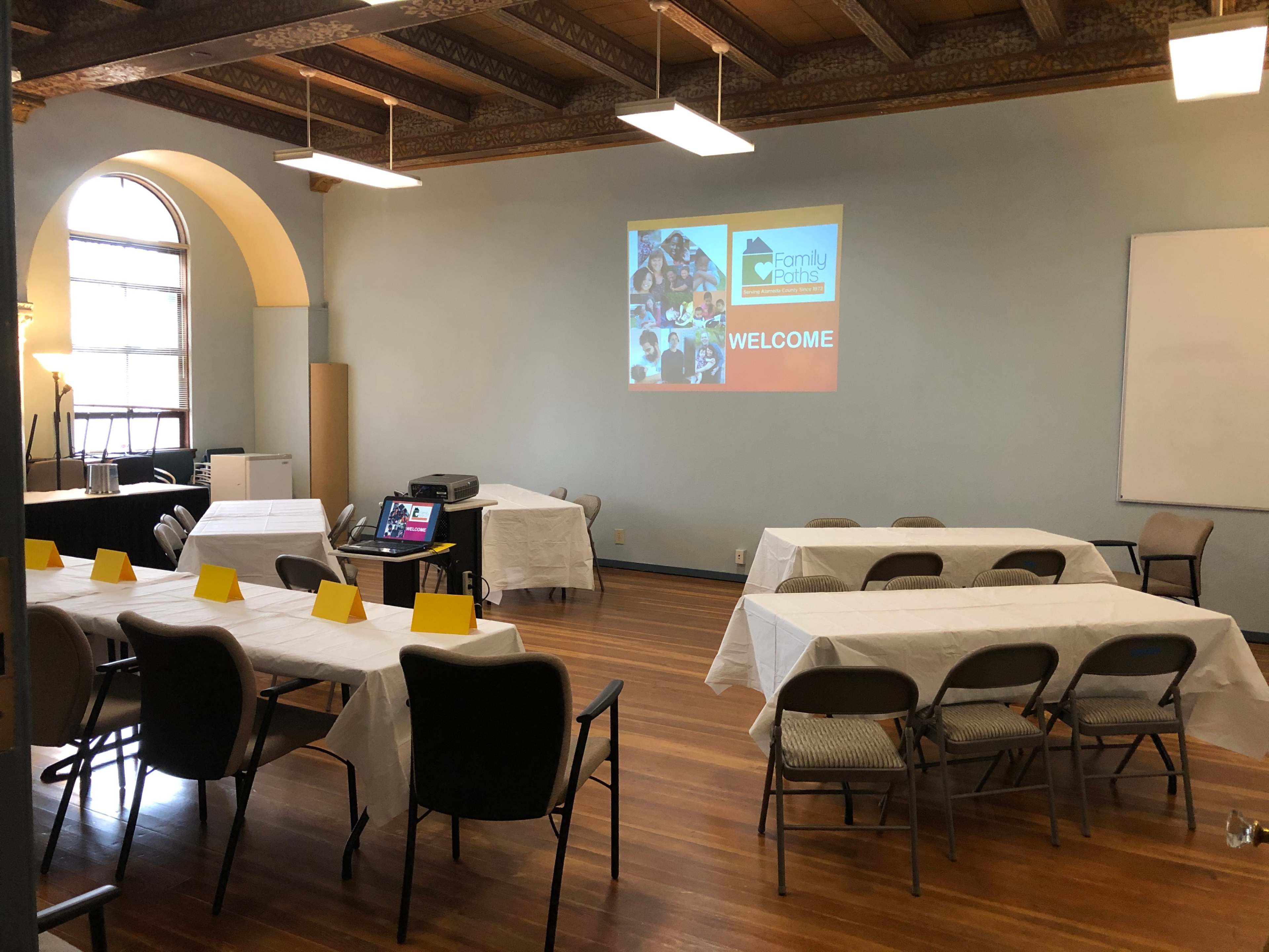 A conference room setup with several tables covered in white tablecloths, chairs arranged around them, and a projector displaying a "WELCOME" message on the wall.