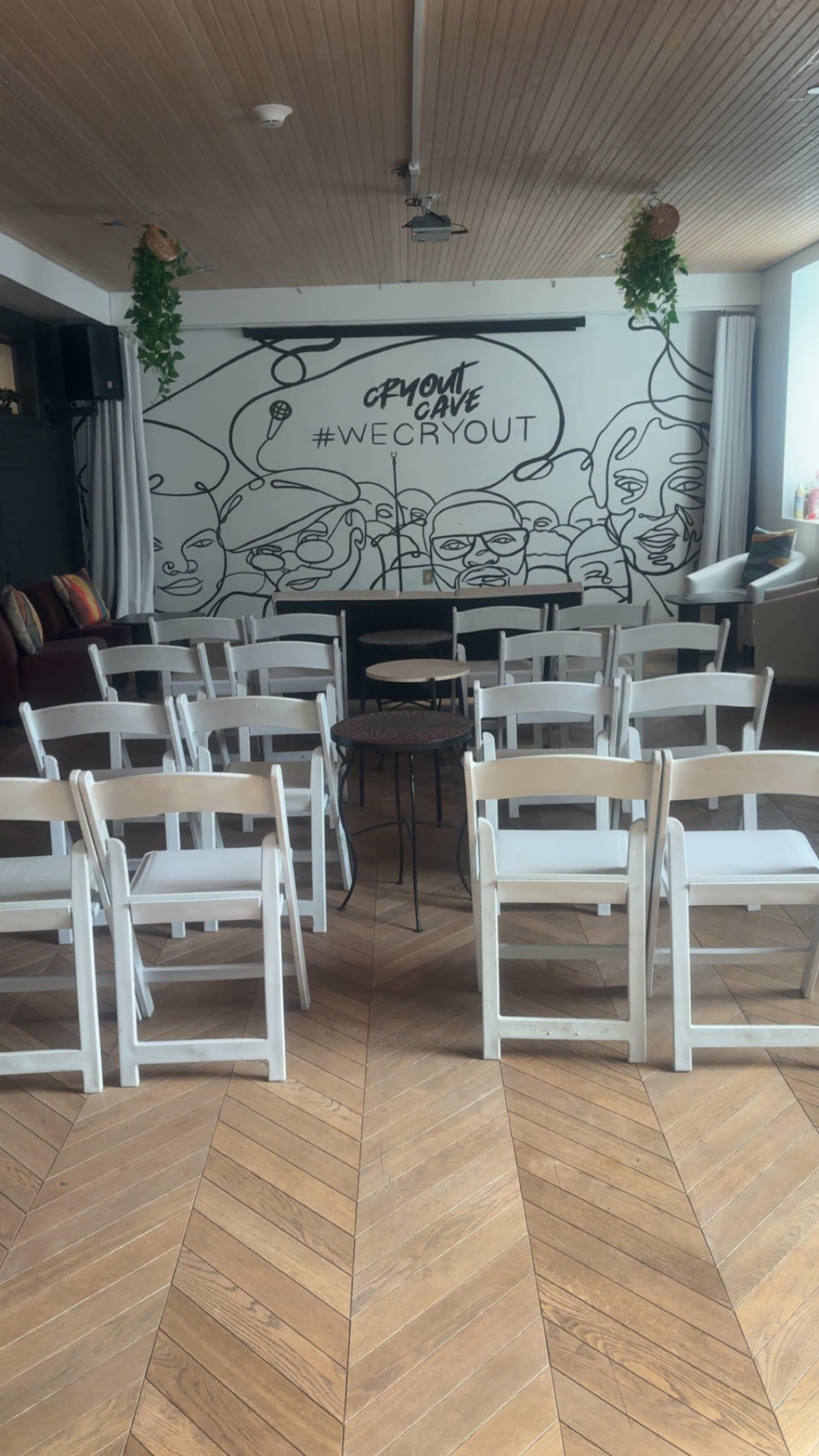 The image shows an empty room set up with rows of white folding chairs facing a small table, with a mural featuring abstract faces and the phrase "#WECRYOUT" on the wall behind them.