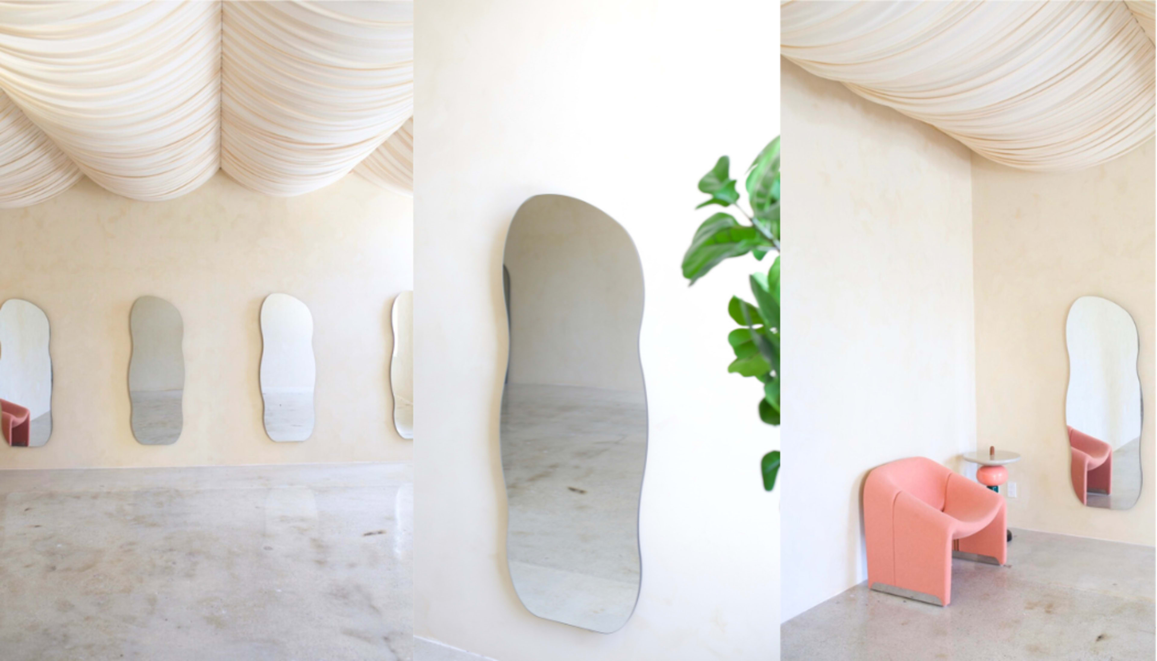 The image shows a minimalist interior space with curved mirrors on one wall, a pink chair, and a small table near a leafy plant, all beneath a draped ceiling.