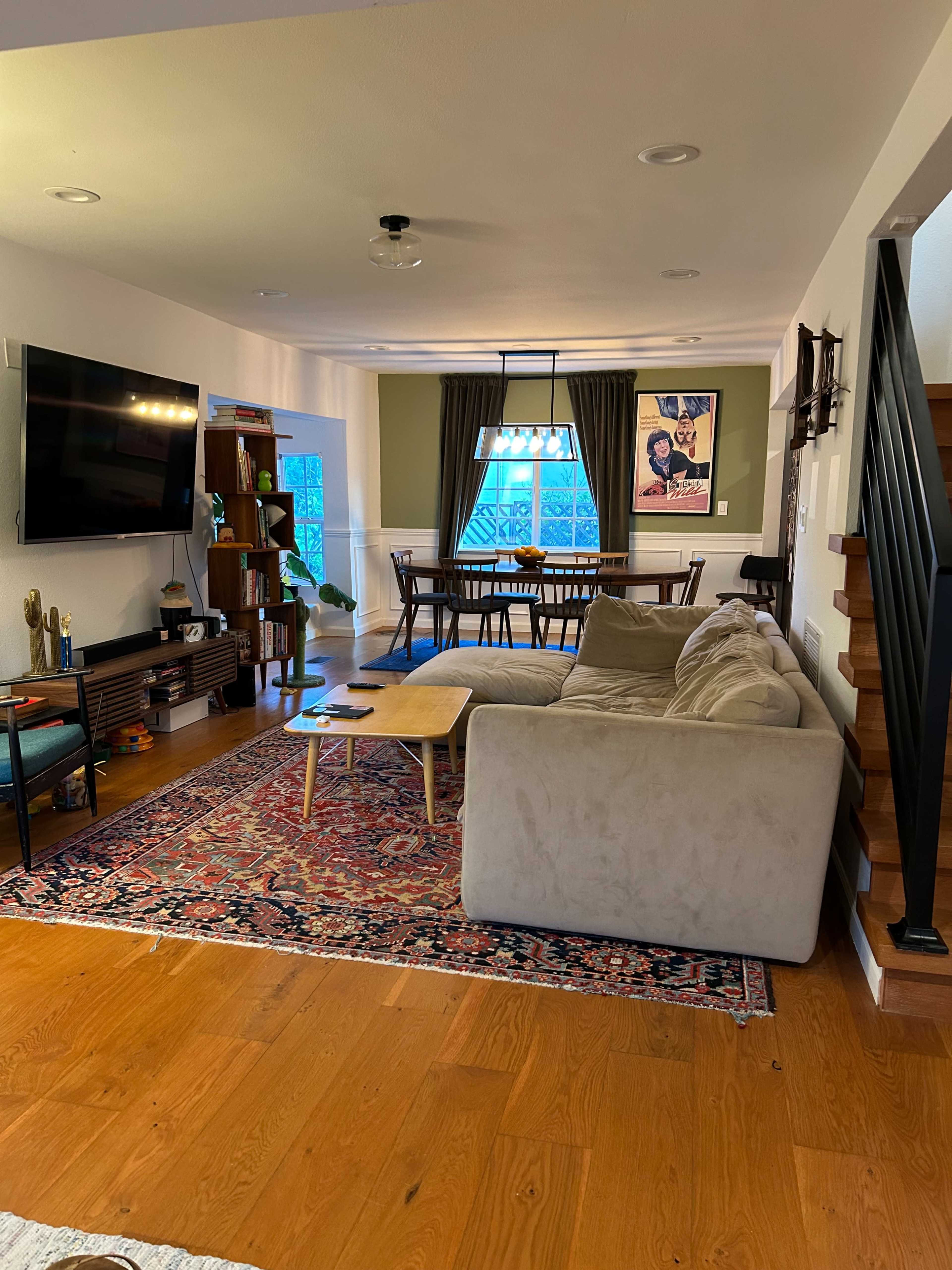 A spacious living room with a beige sectional sofa, a large television mounted on the wall, and a wooden dining table set against green walls.