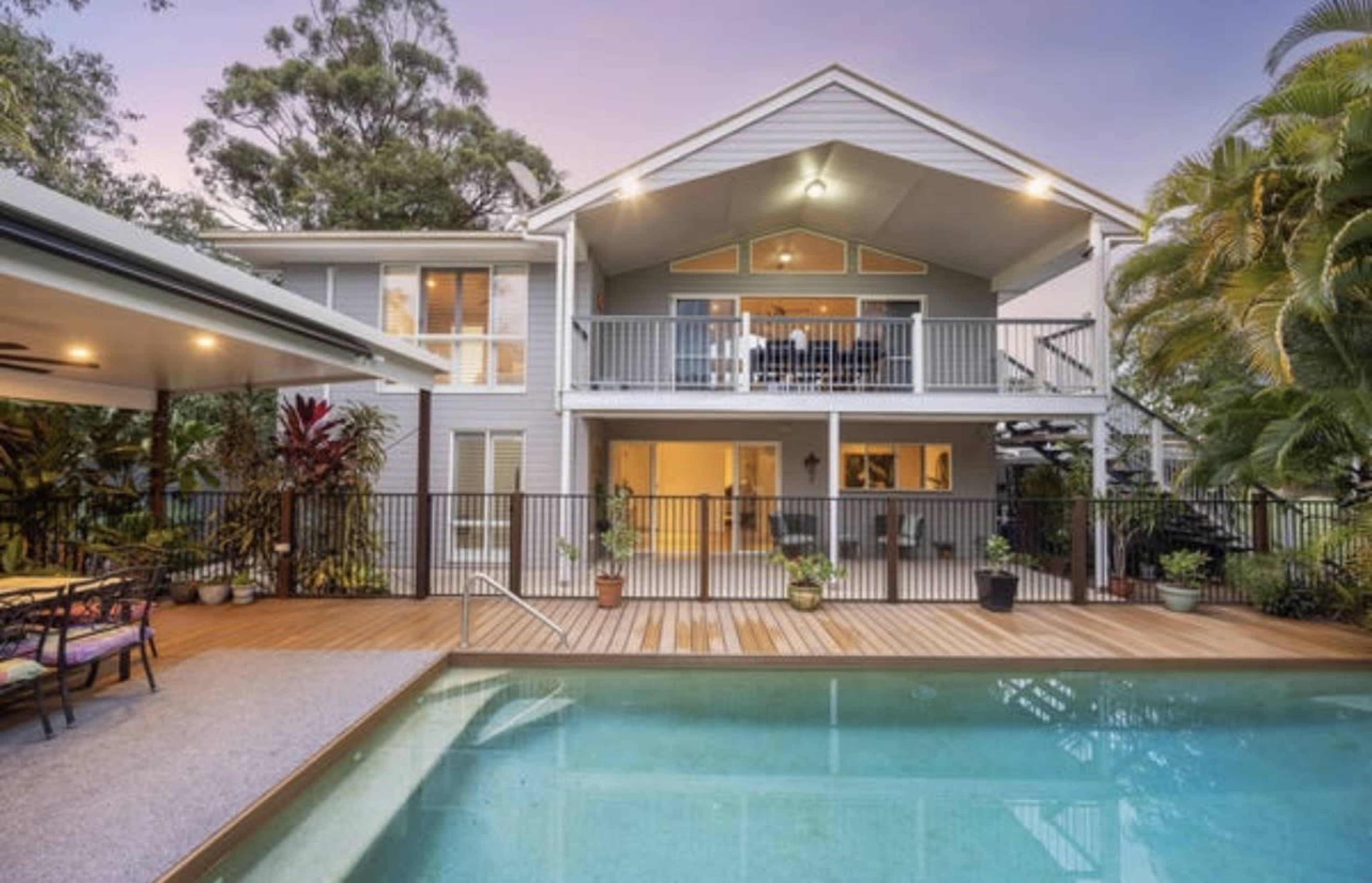 The image shows a modern two-story house with a balcony, surrounded by lush greenery and featuring a swimming pool in the foreground.
