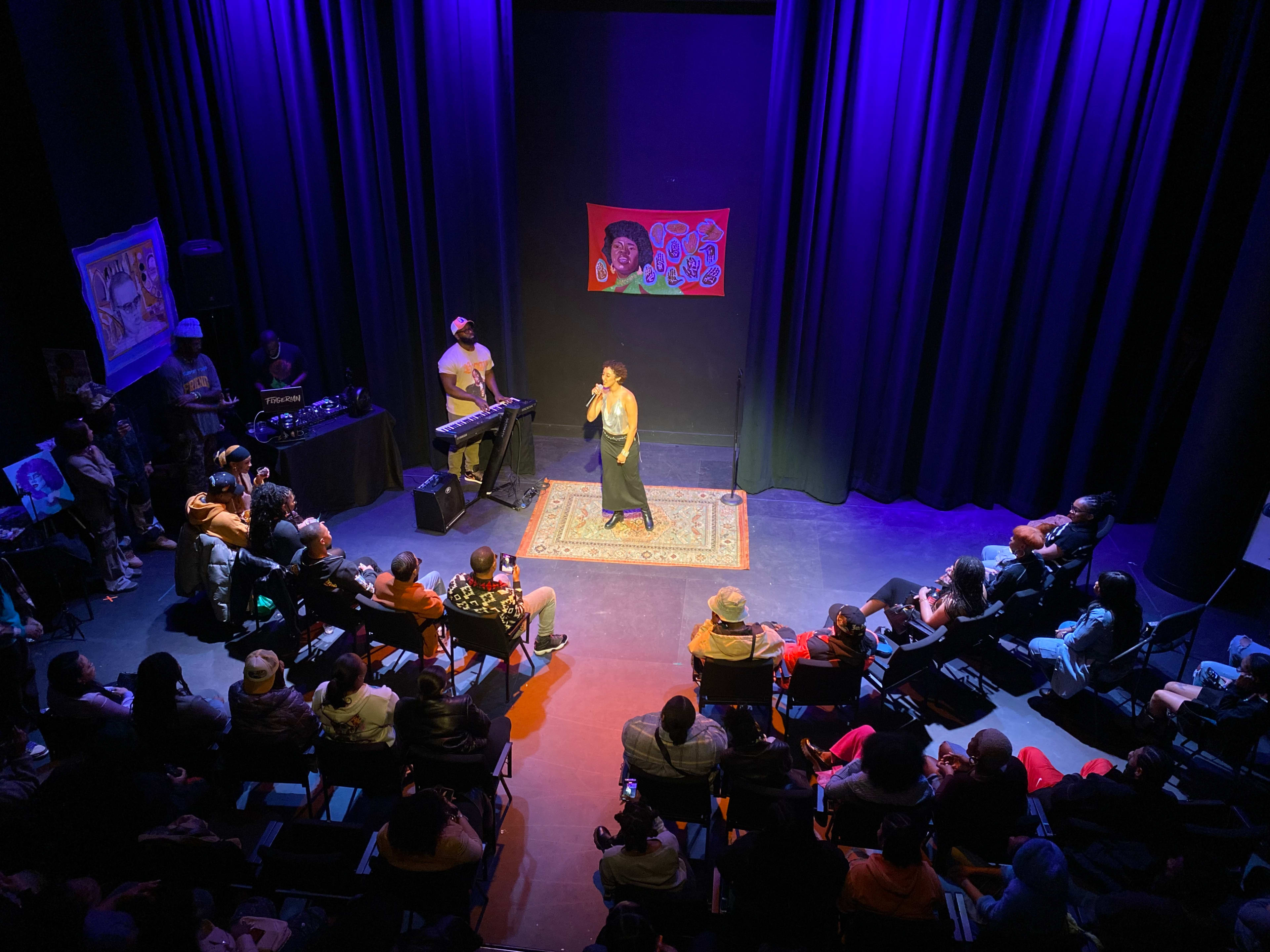 A performer stands on stage in front of an audience beneath purple lighting, with a keyboardist nearby and artwork displayed on the walls.