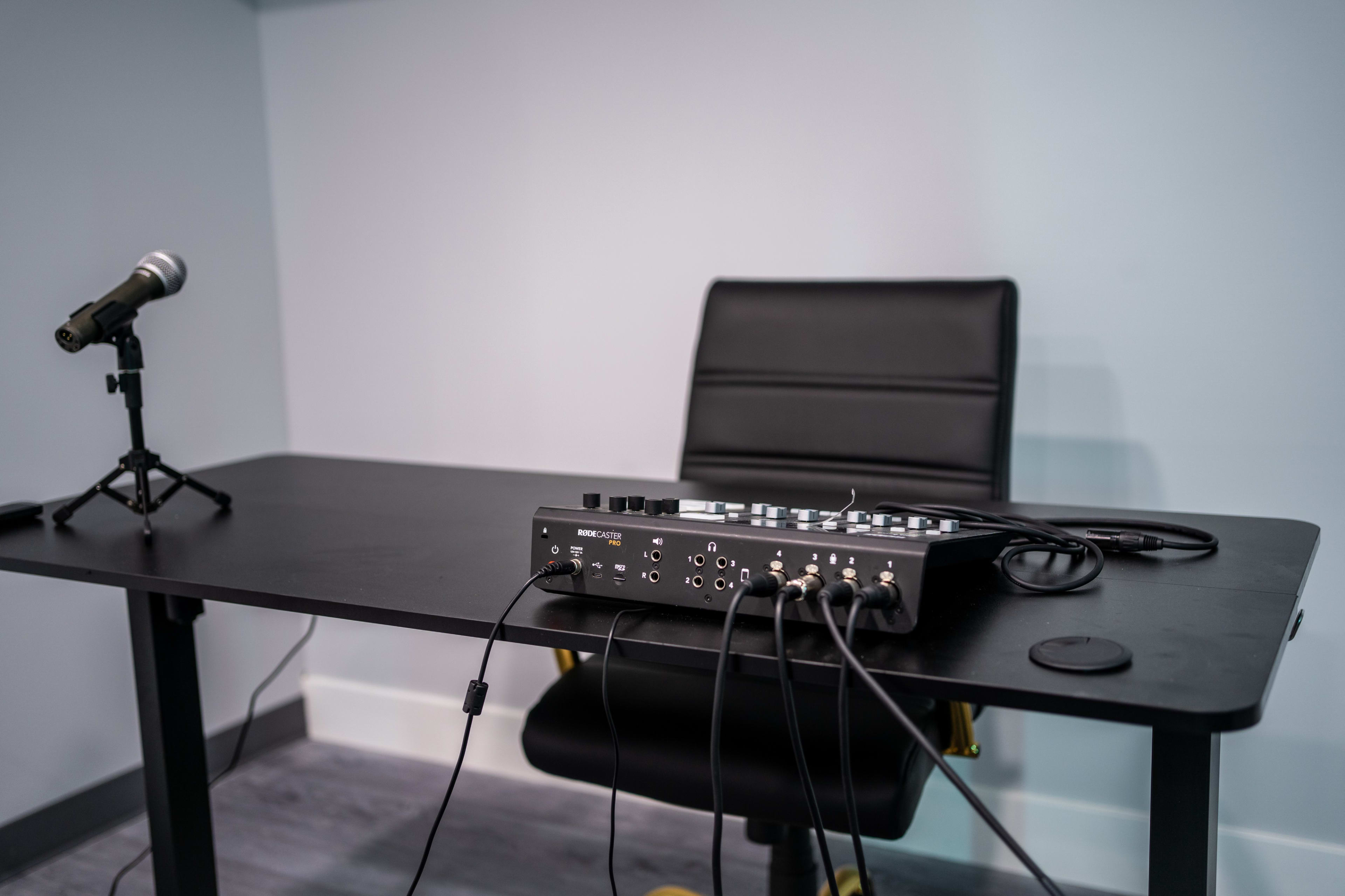 A black desk with a microphone stand and audio mixer sits against a light blue wall.