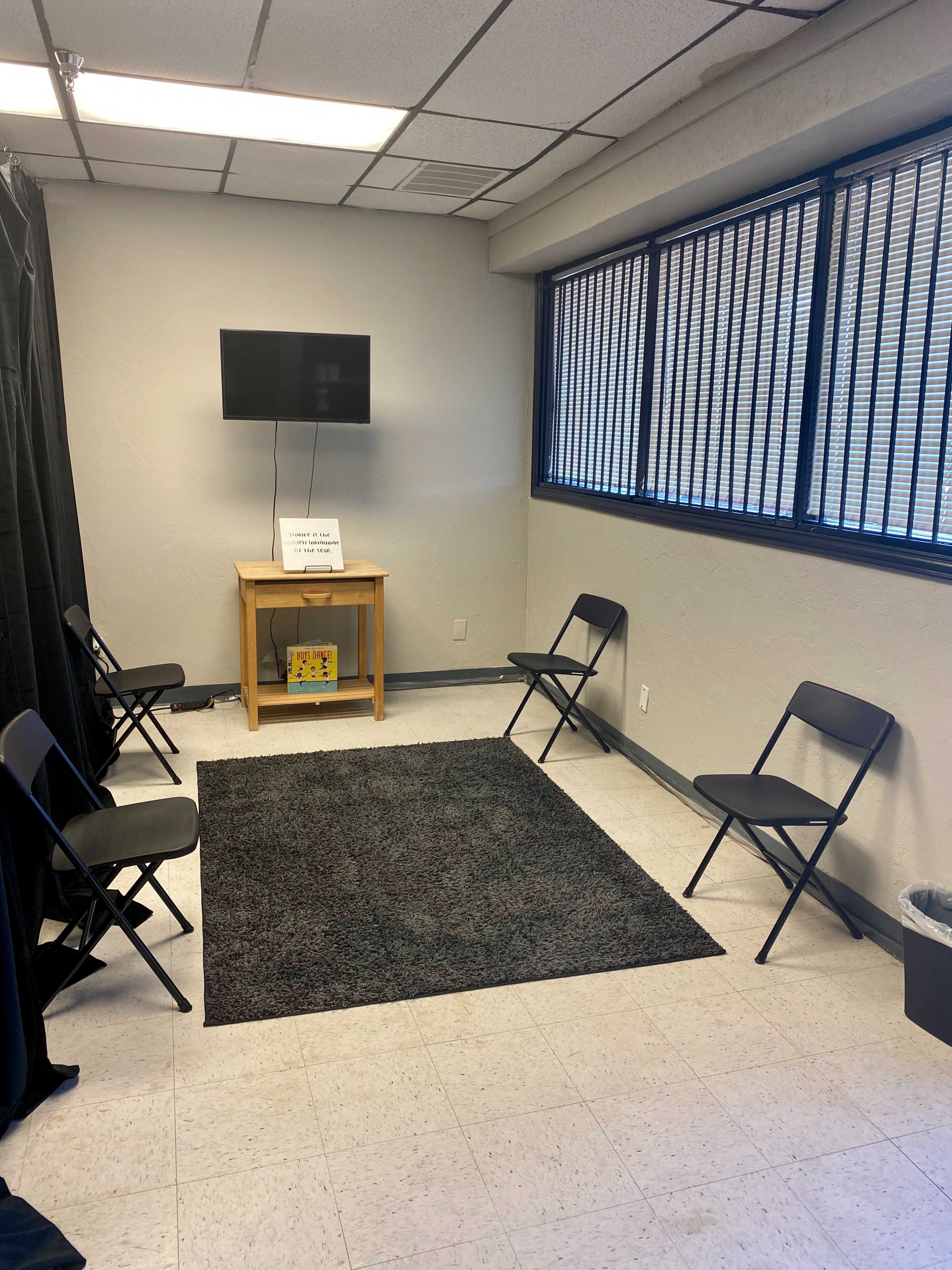 A small waiting room features a gray rug on the floor with four black chairs arranged around it, a wooden table holding a TV, and large windows with blinds.