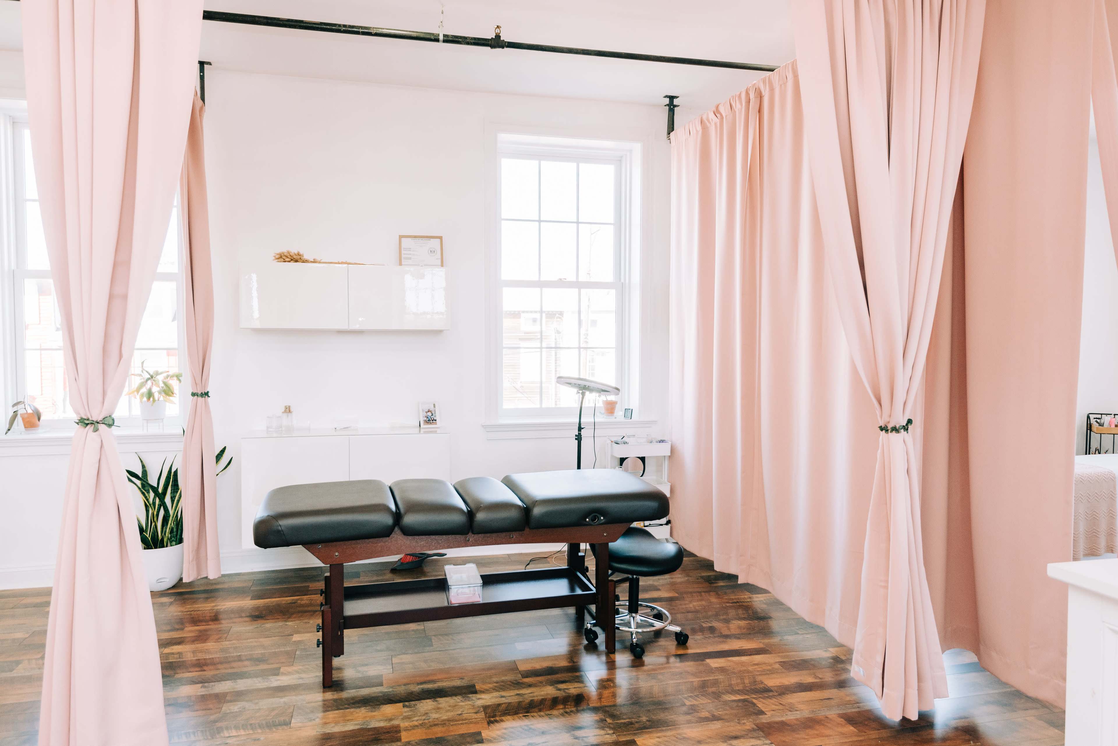 The image shows a bright therapy room with a massage table, a rolling stool, and light pink curtains separating the space.