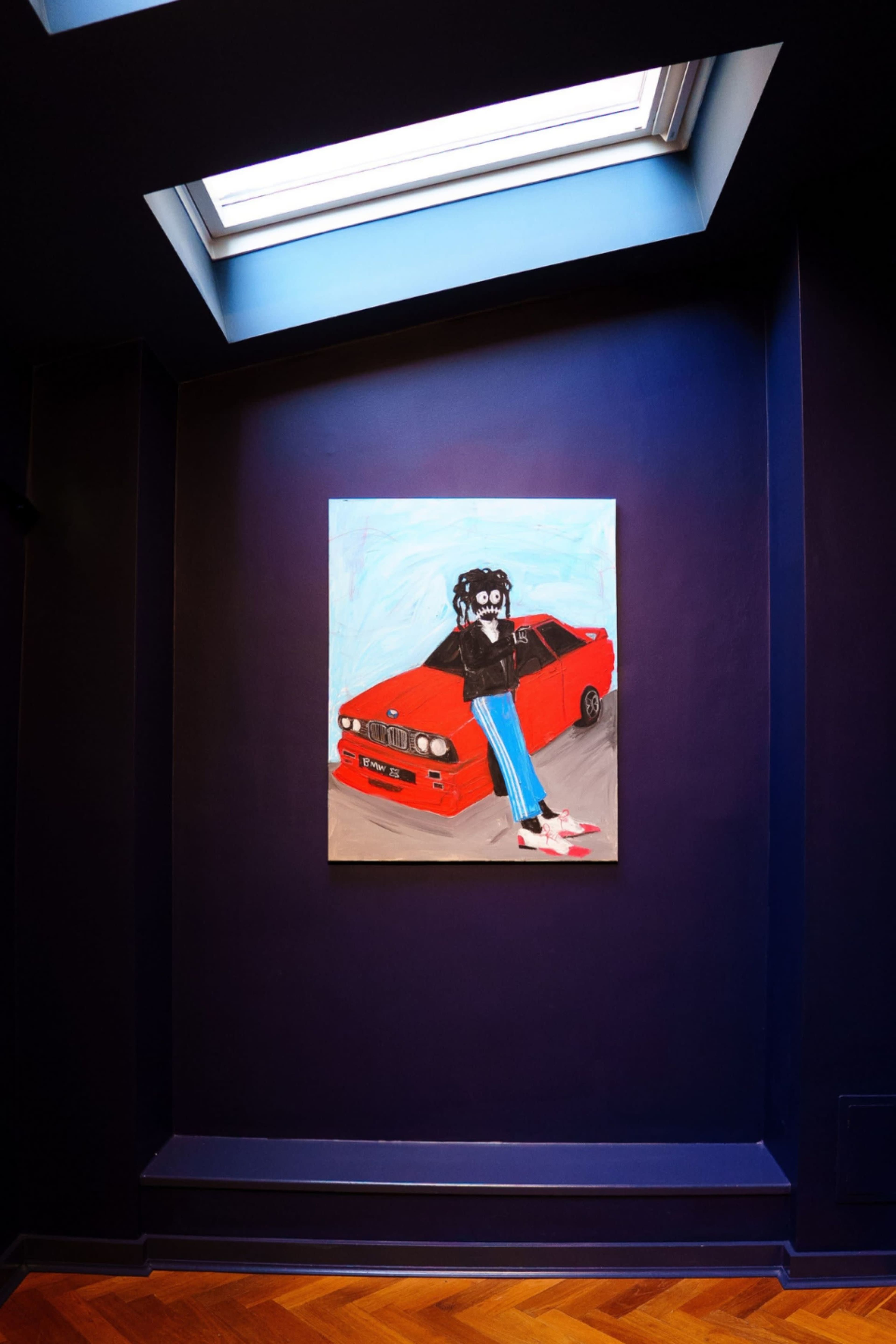 The image shows a colorful painting of a figure standing on a red car, displayed on a dark blue wall beneath a skylight.