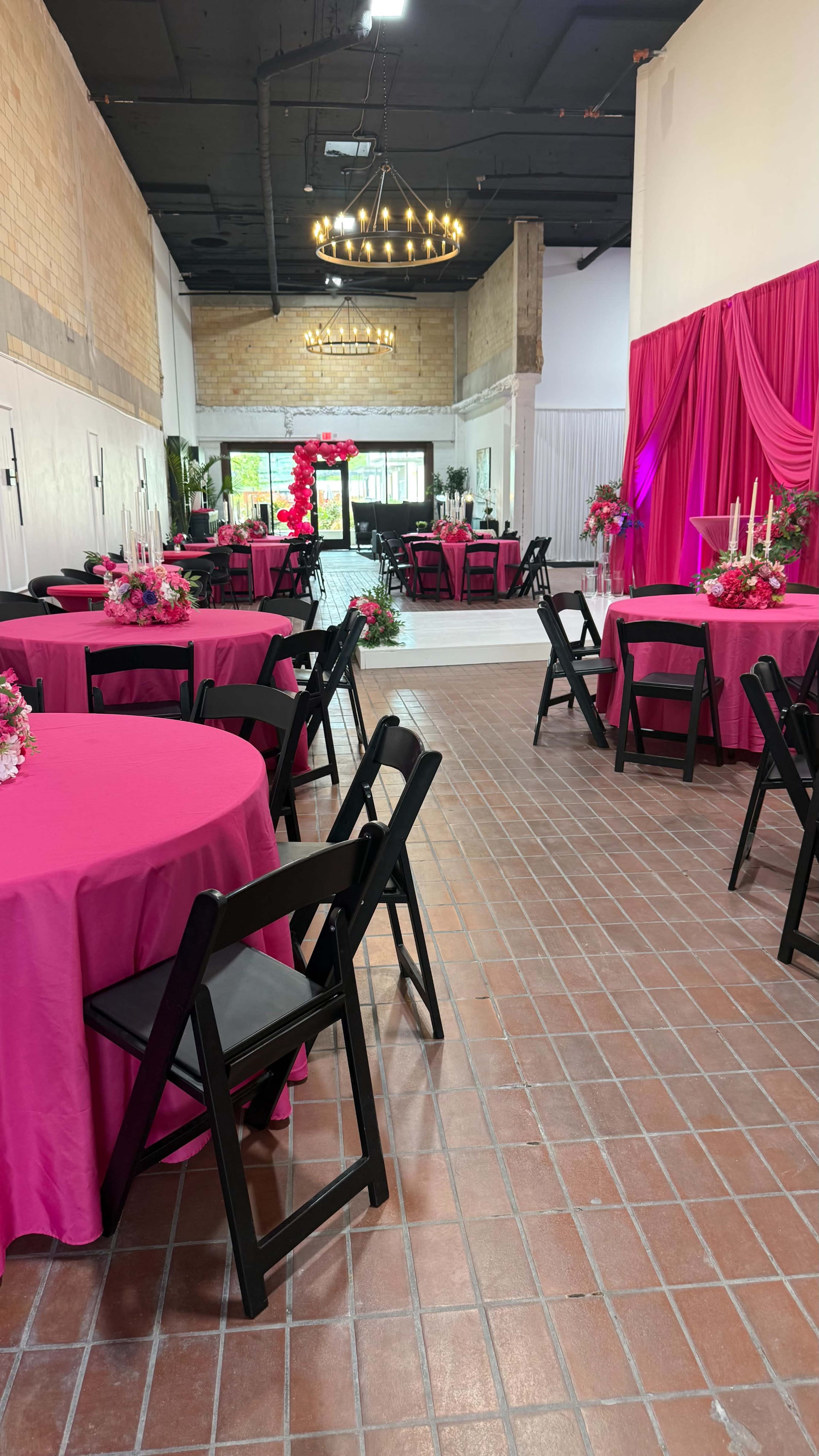 The image shows a decorated event space with pink tablecloths on round tables, black folding chairs, and floral centerpieces, illuminated by a chandelier overhead.