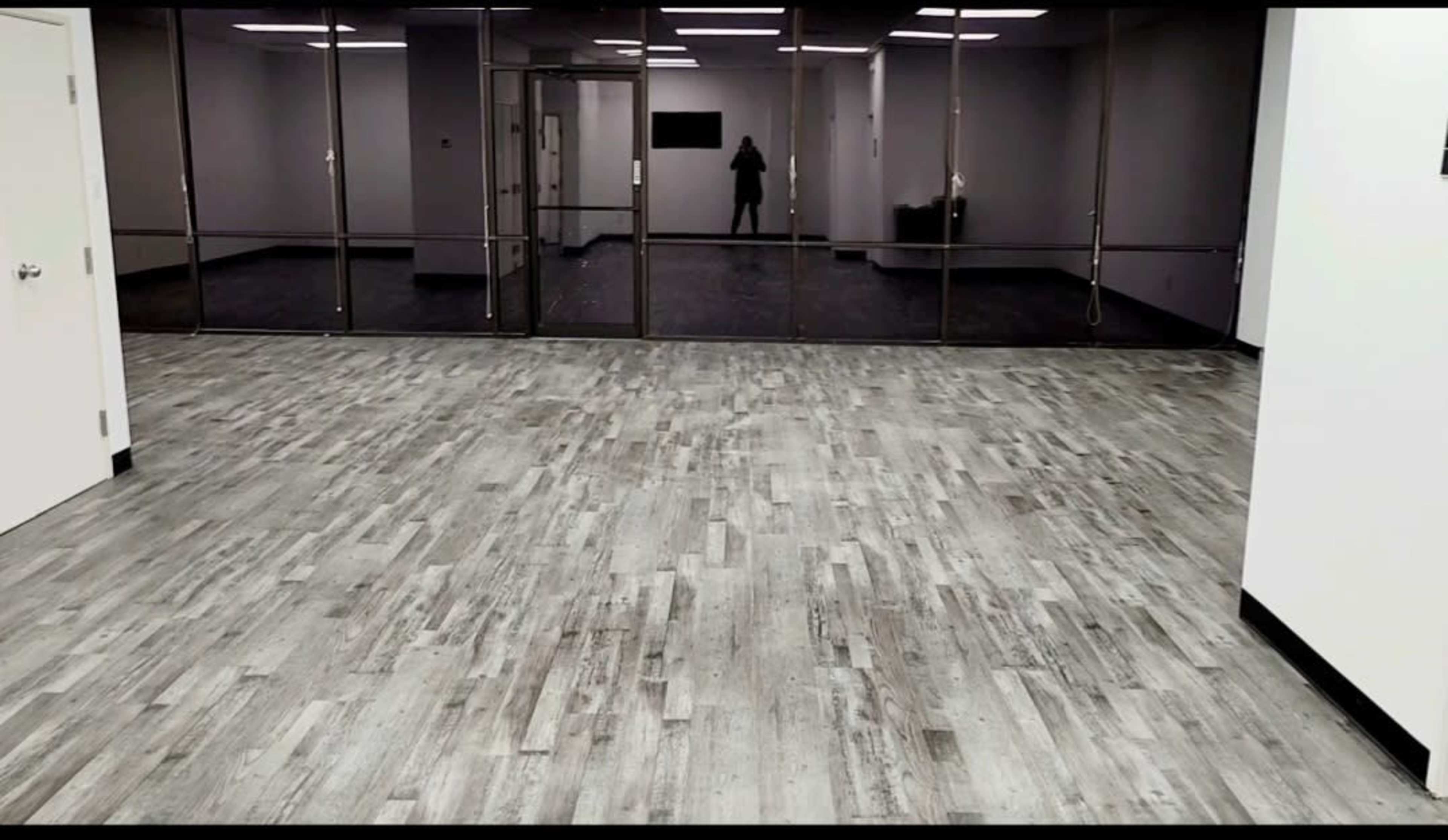 An empty room with a wooden floor and large glass windows reflecting a figure in the background.