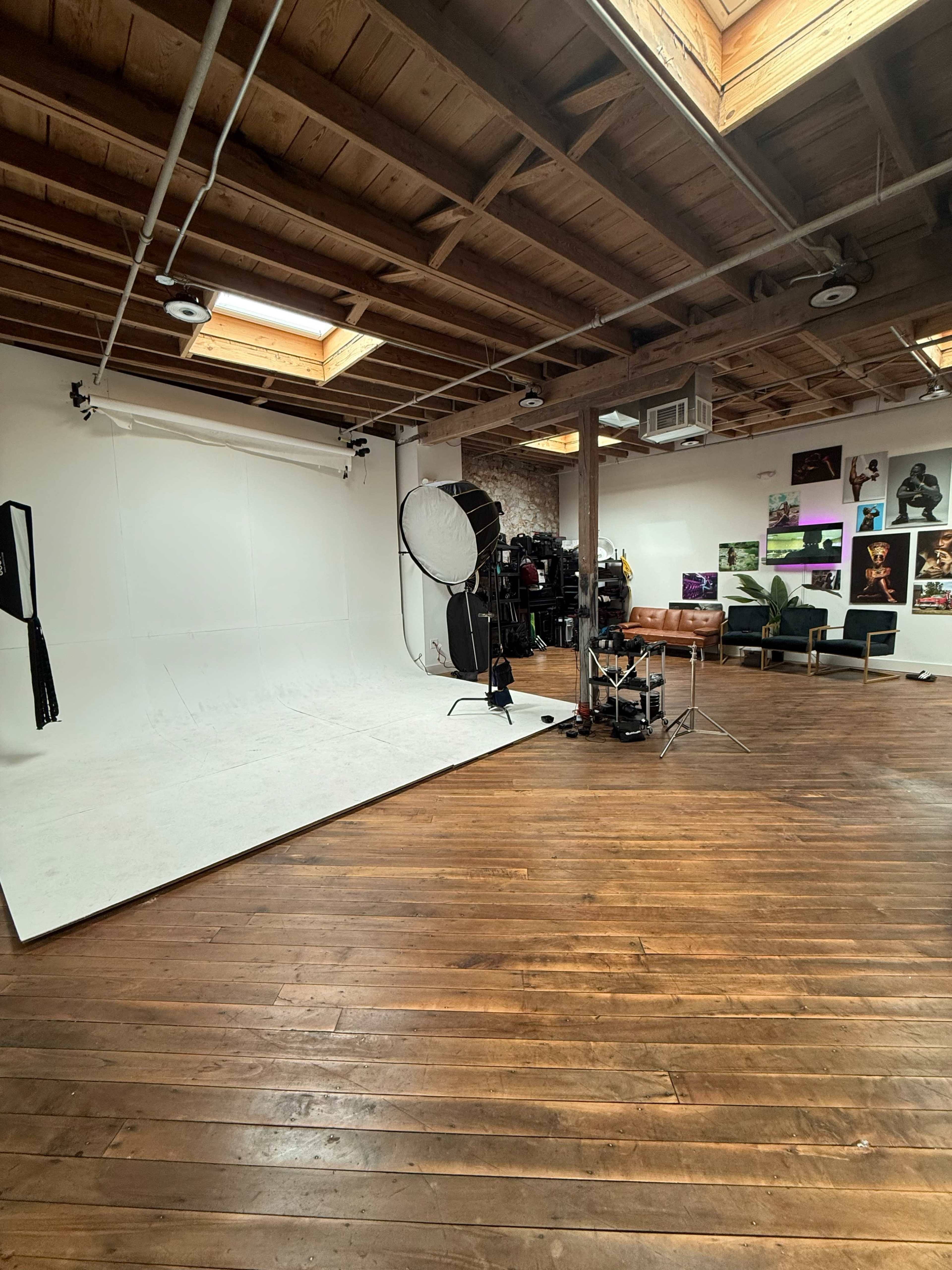 The image shows a photography studio with a white backdrop, lighting equipment, and various seating arrangements against wooden floors.