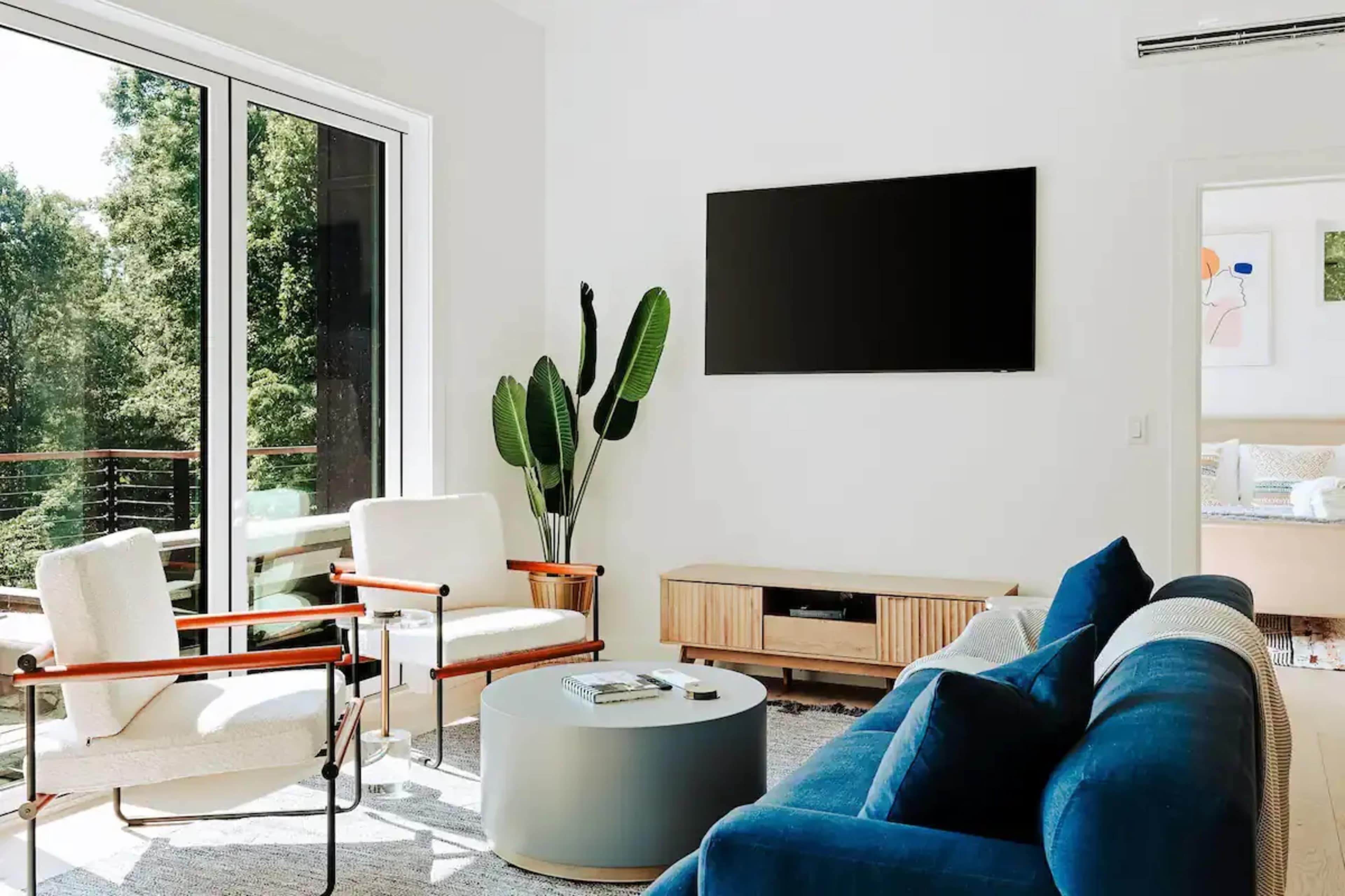 A modern living room with a blue sofa, two white chairs, a circular coffee table, and a television mounted on the wall, surrounded by large windows and a green plant.