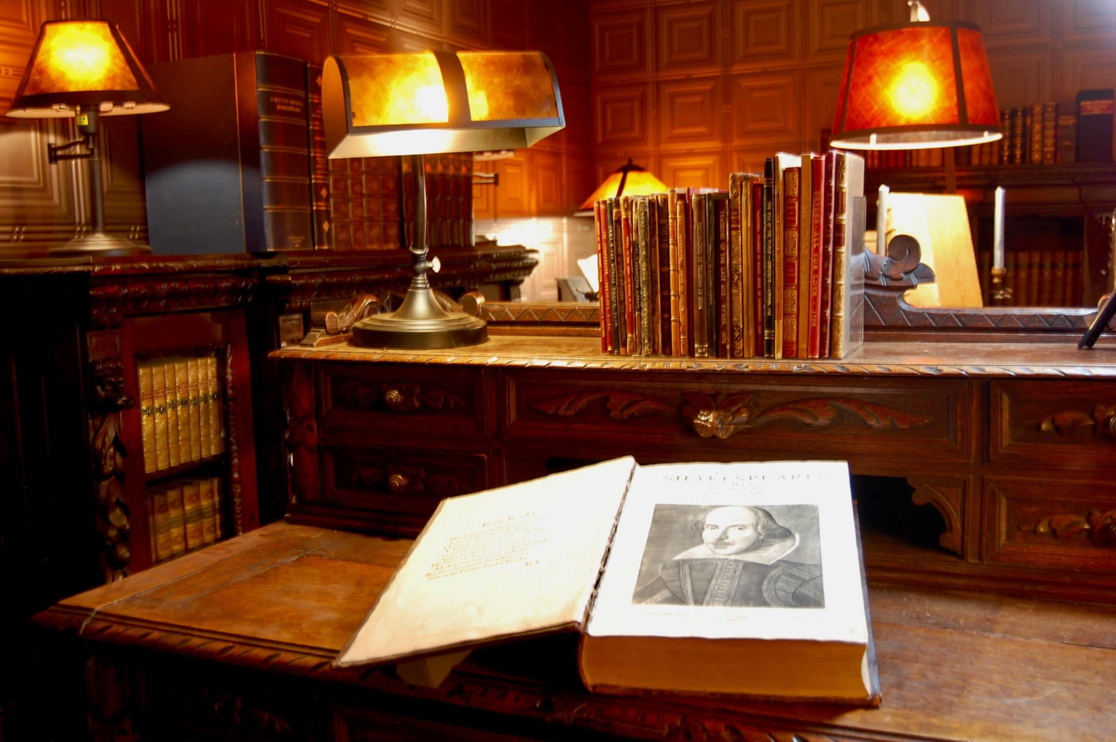 A wooden desk is adorned with books and lamps, featuring an open book displaying an engraving of a historical figure, set against a backdrop of shelves filled with additional volumes.