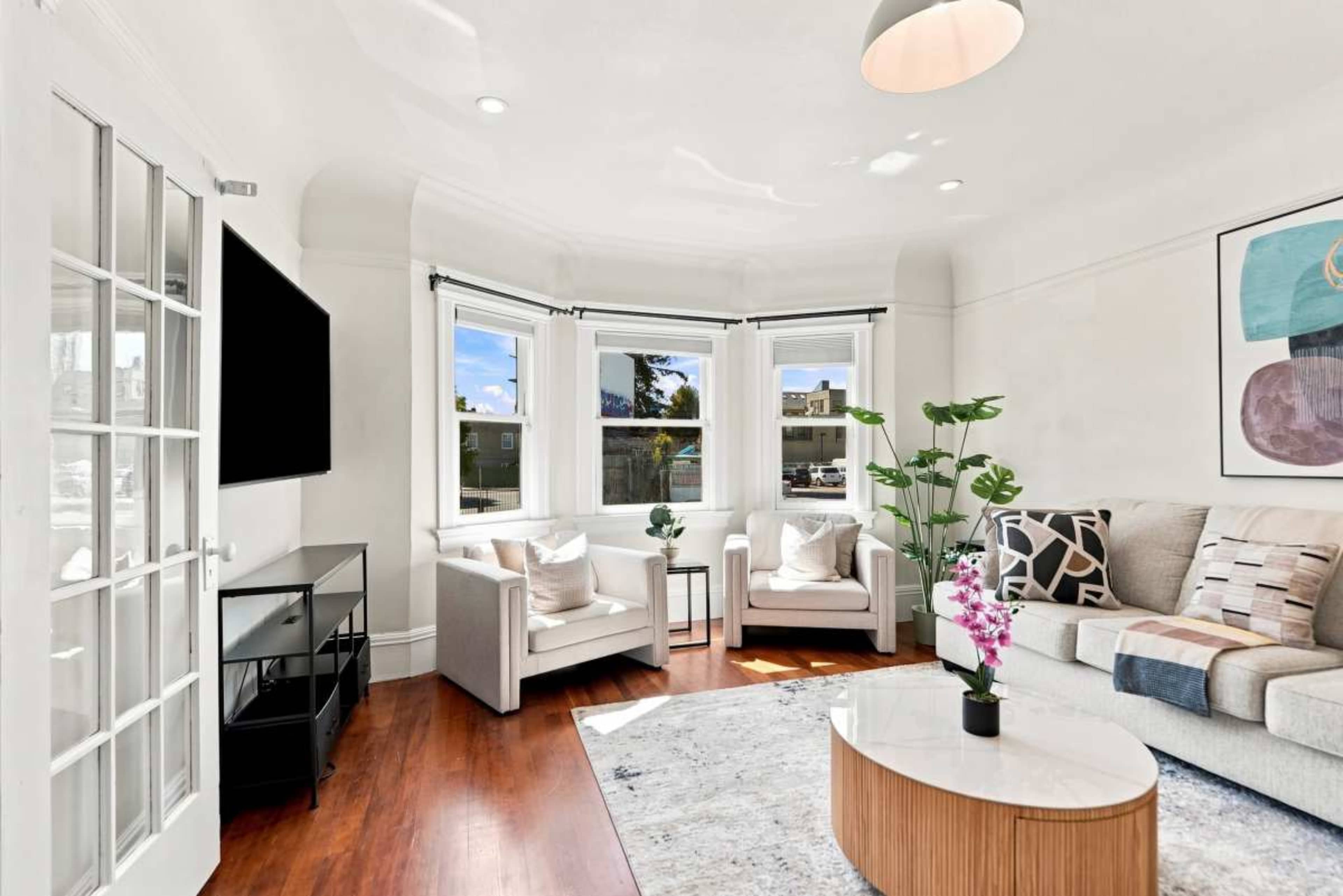 A bright living room features a sectional sofa, two armchairs, a round coffee table, a flat-screen TV mounted on the wall, and large windows allowing natural light to enter.