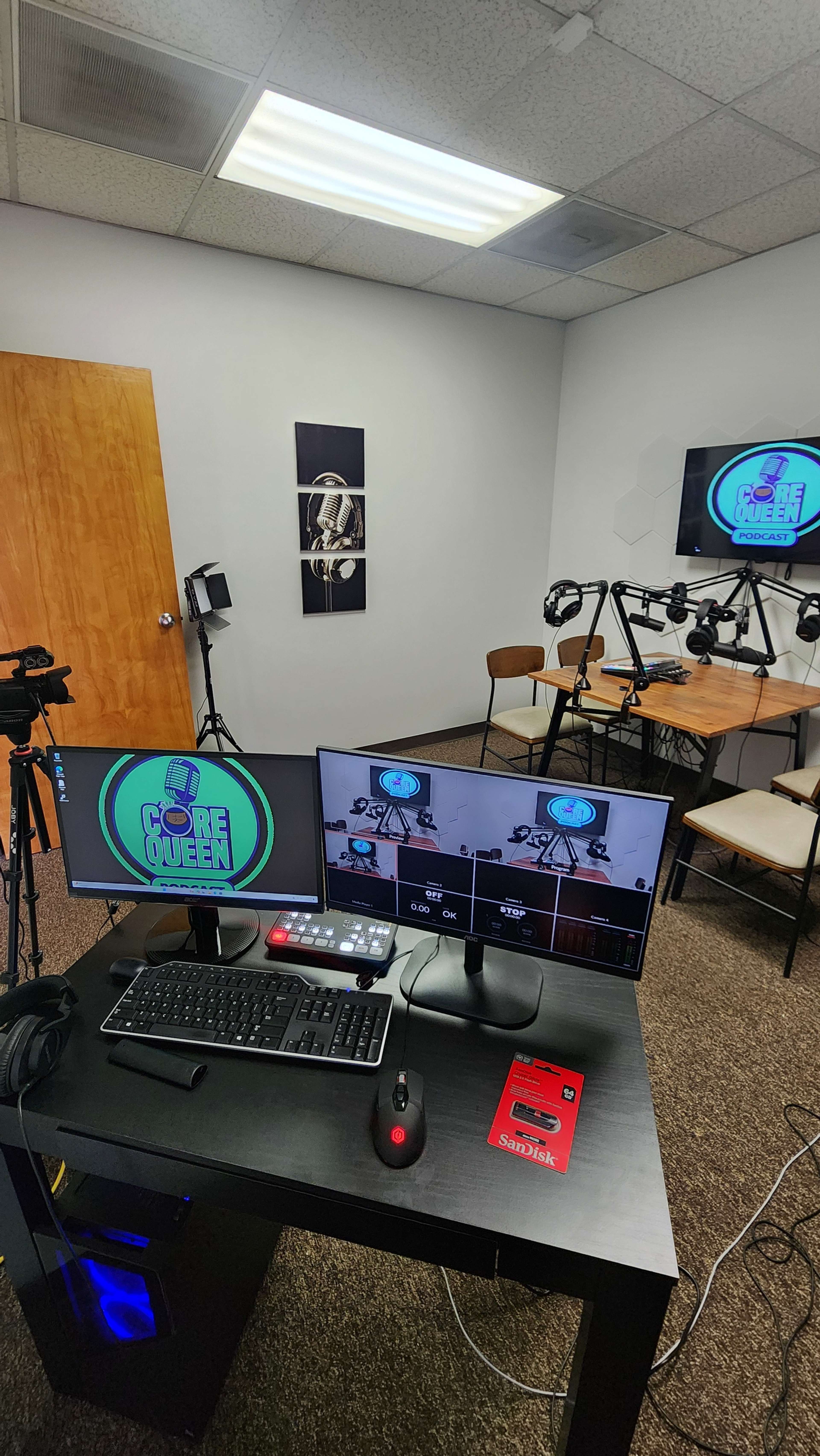 A media production setup with two computer monitors displaying video feeds, a keyboard, cameras, and a microphone, all positioned in a small room with a wooden door and wall artwork.